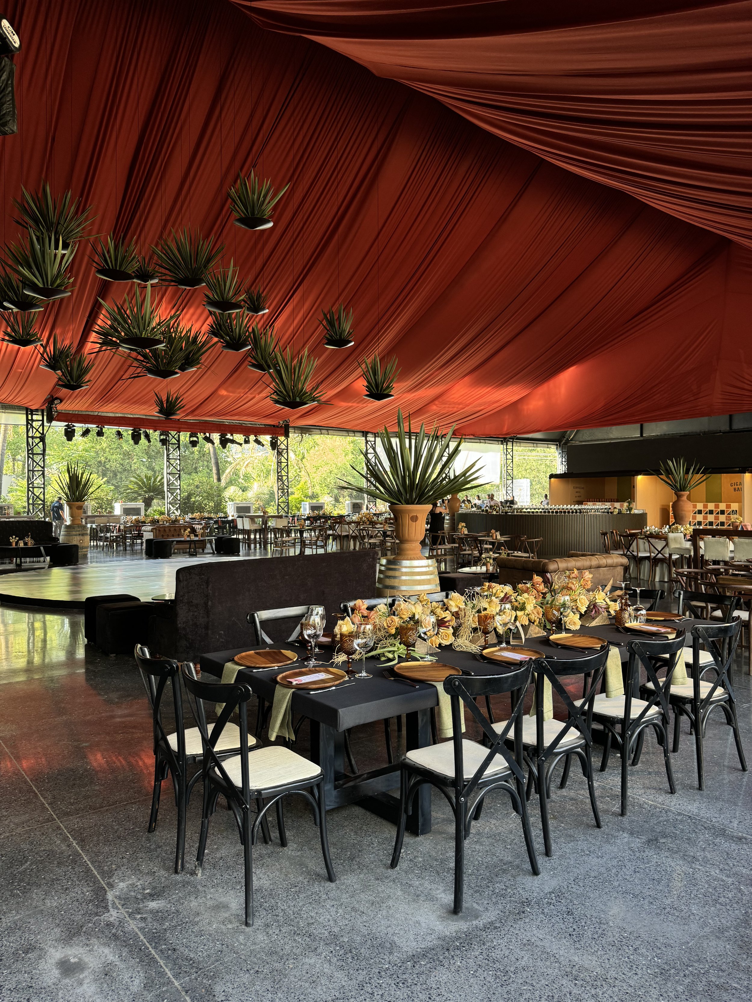 Elegant dining area with a long black table decorated with a floral centerpiece, surrounded by black chairs with beige cushions, under a red ceiling with hanging plants.