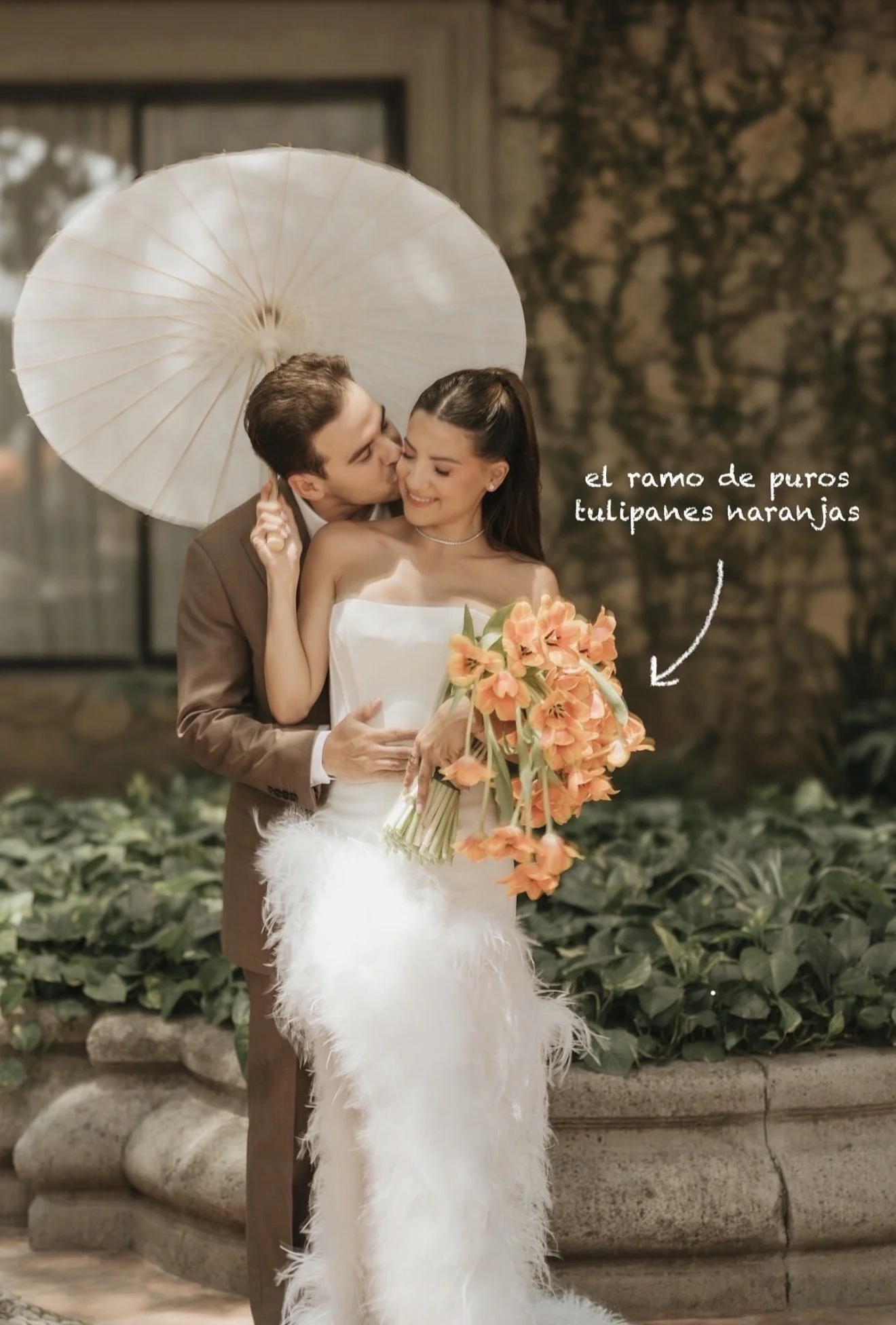 A couple dressed in wedding attire stands outdoors, sharing an intimate moment with the groom kissing the bride's cheek. The bride wears a strapless white wedding dress with feather details at the bottom and holds a bouquet of orange narcissus flower