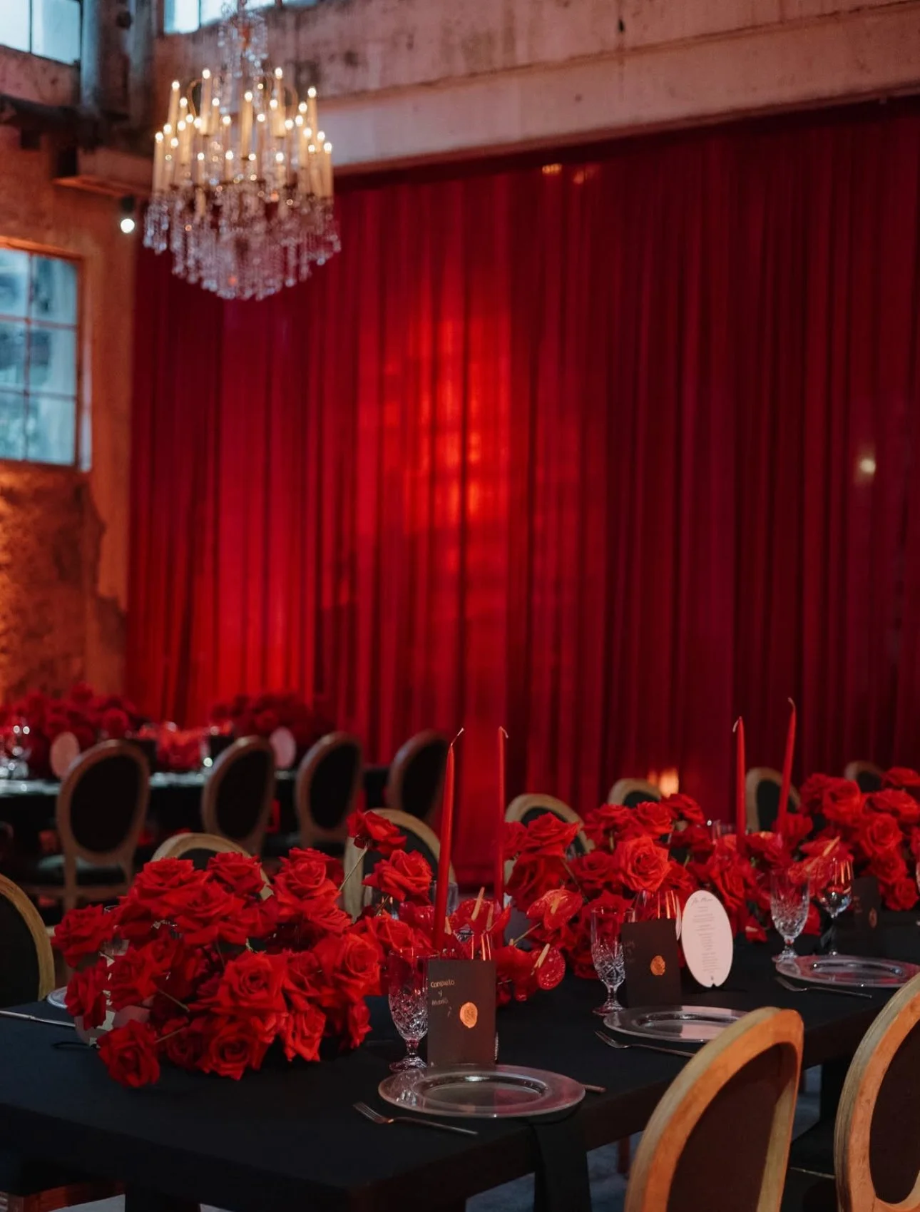 A banquet table decorated with red roses, candles, and glassware inside a rustic venue with a red curtain and chandelier.
