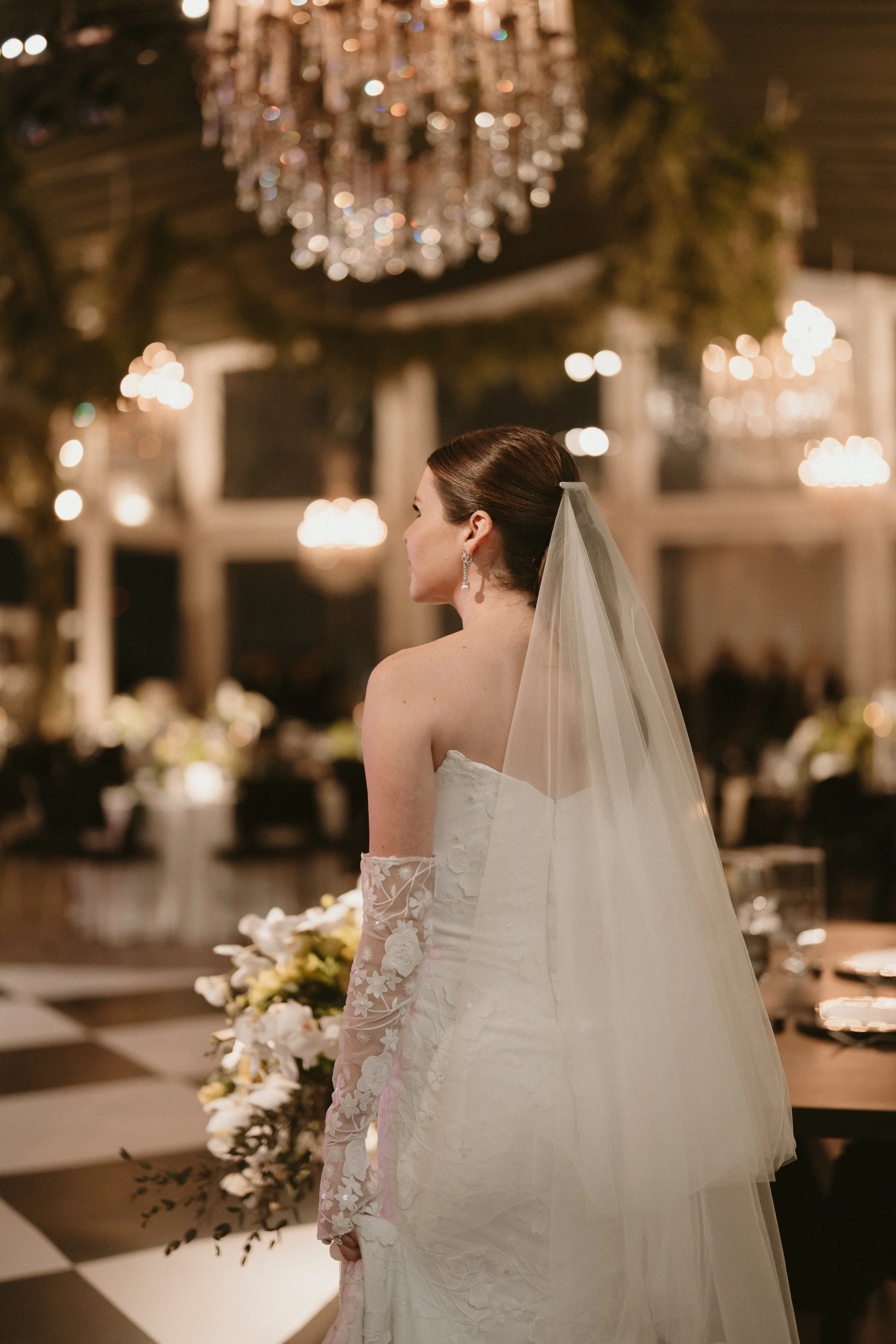Bride in a strapless wedding gown with sheer embroidered long sleeves, wearing earrings and a veil, standing in an elegant decorated venue with chandeliers and floral arrangements.