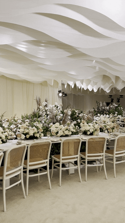 Elegant event space with a long table decorated with white and blush flowers, surrounded by chairs, under a white, wavy fabric ceiling.