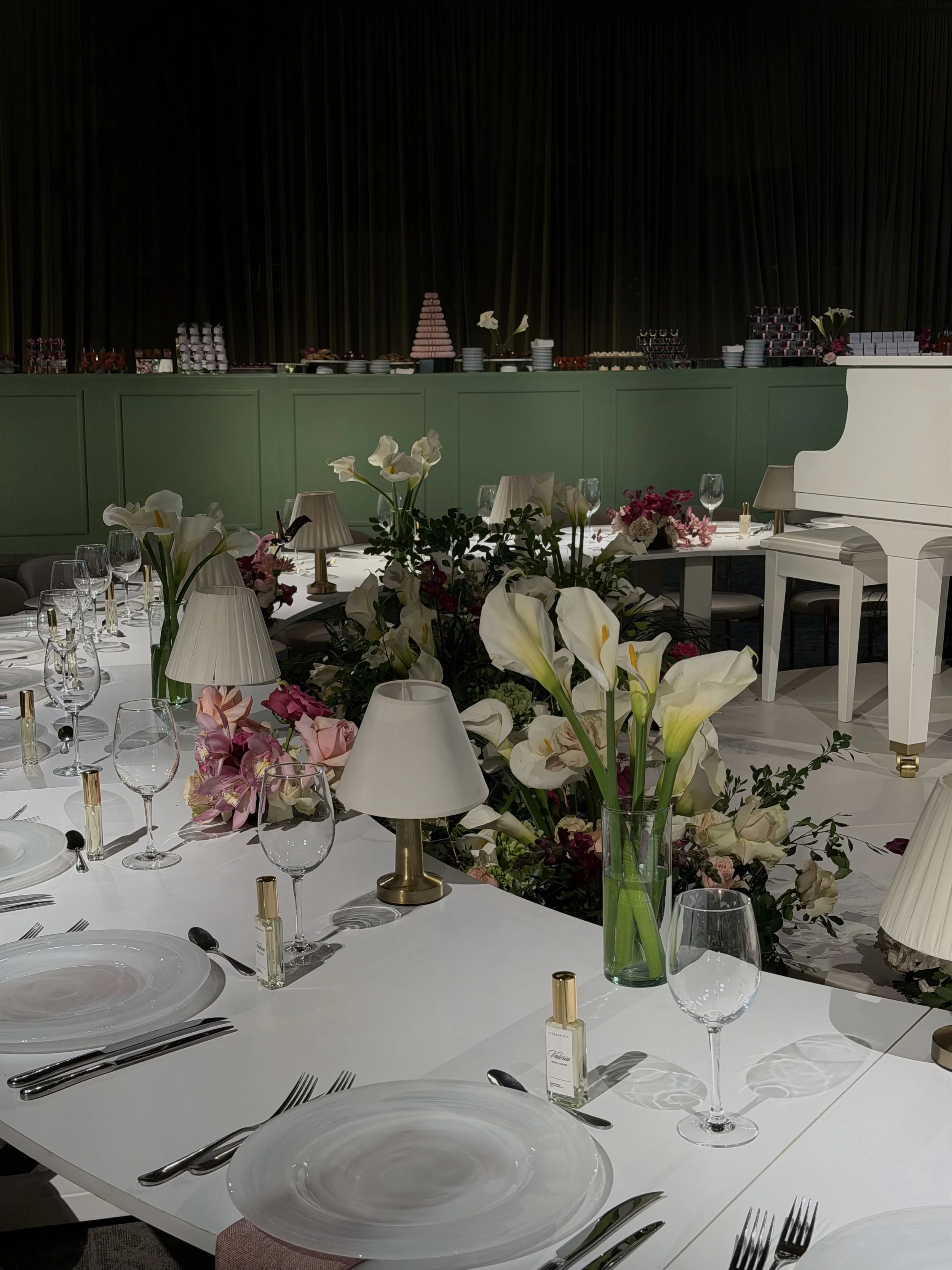 Elegant banquet table with white tablecloth, floral centerpiece, lamps, and place settings, in a decorated event space with a green and black backdrop, and desserts on a raised table in the background.
