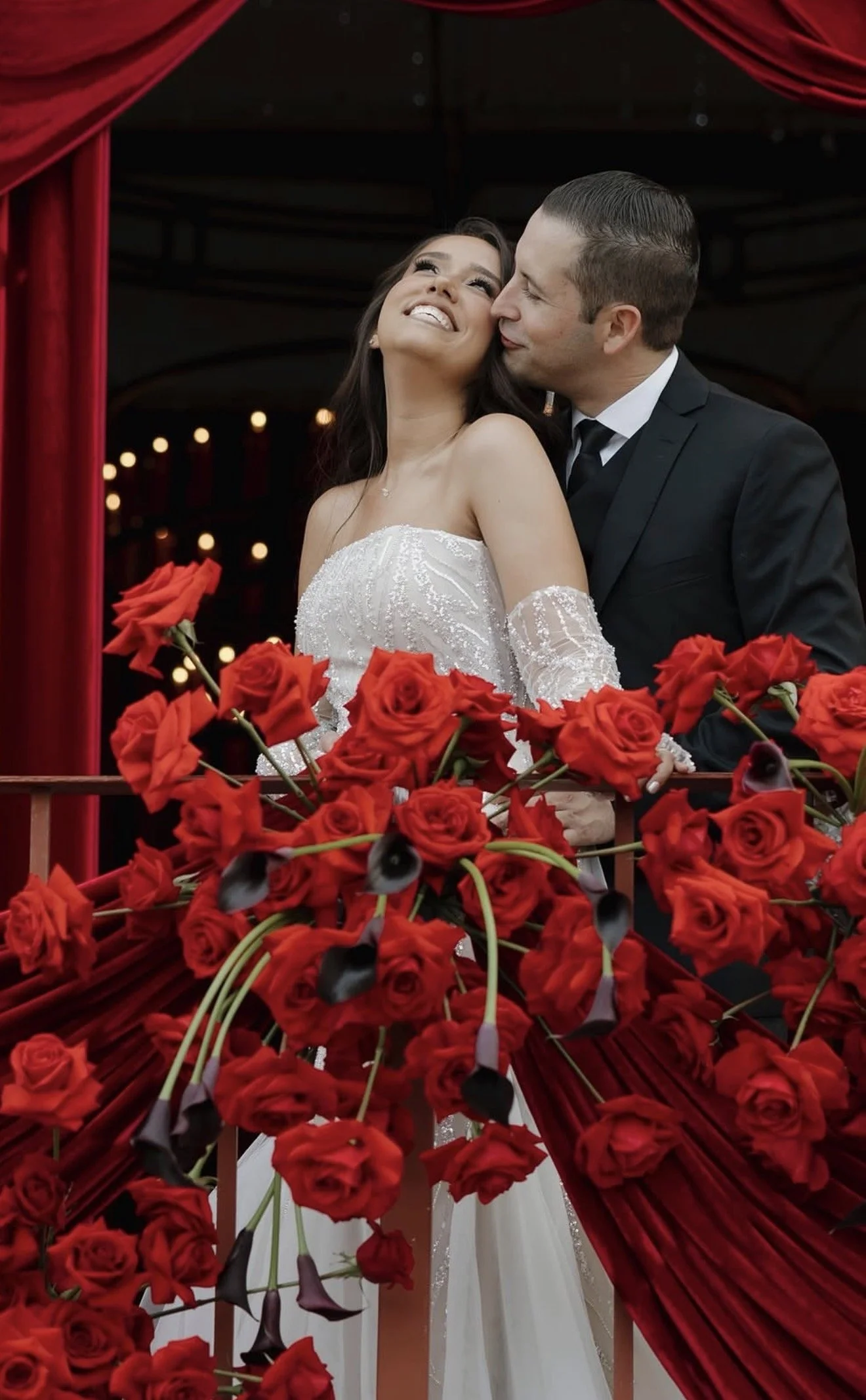 A bride and groom at their wedding, standing on a balcony decorated with red roses and drapery, with warm lights in the background.