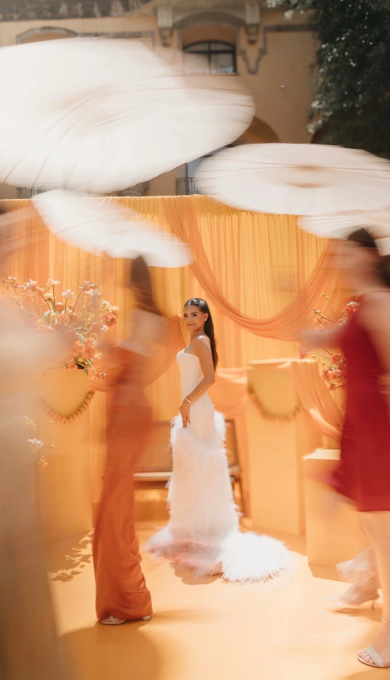 A woman in a white wedding dress stands against an orange backdrop, surrounded by blurred figures and large white decorative clouds, at a festive event or celebration.