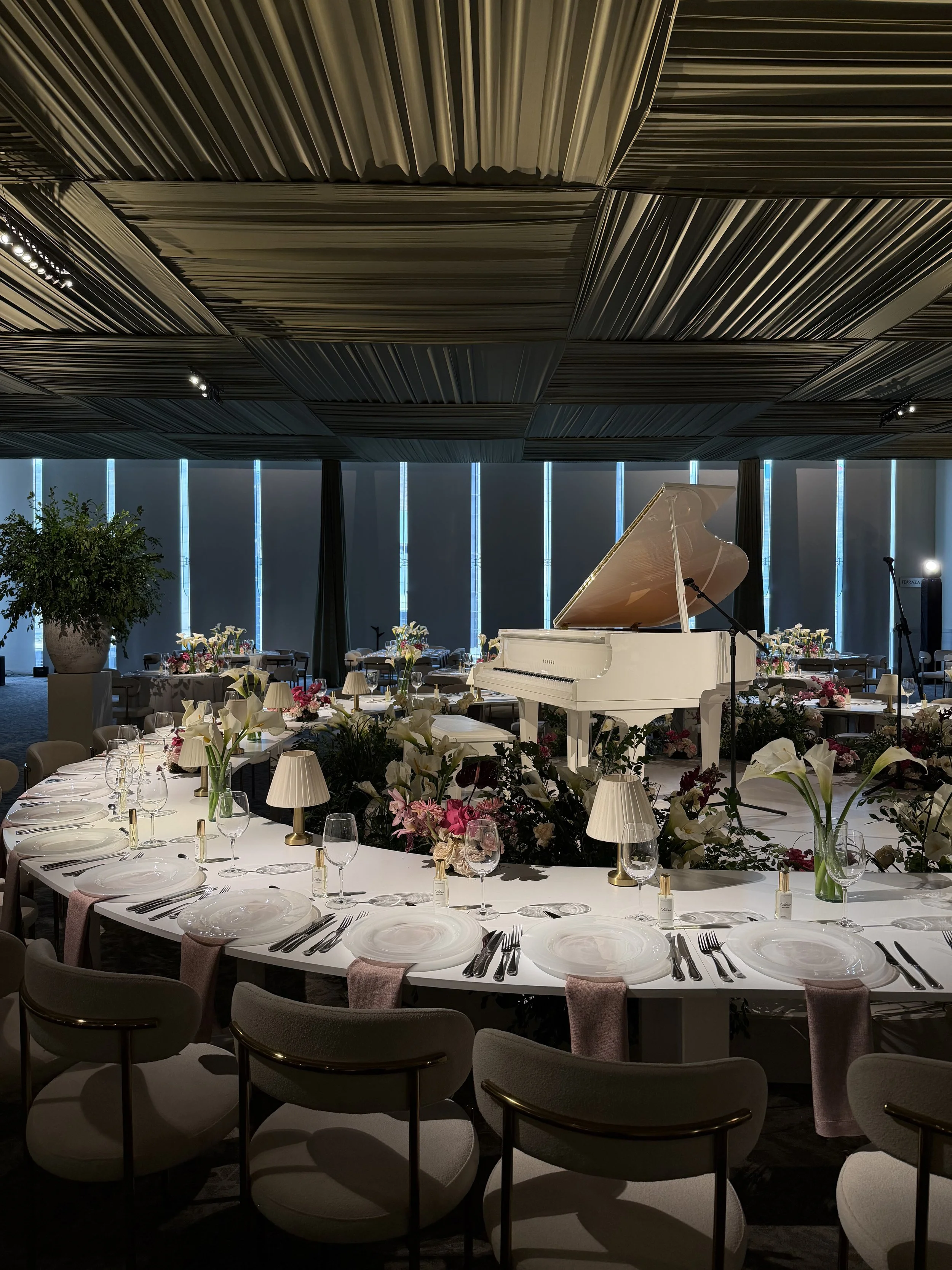 Elegant banquet hall with a white grand piano, surrounded by floral arrangements, table settings, and small lamps, with large windows and a decorative ceiling.