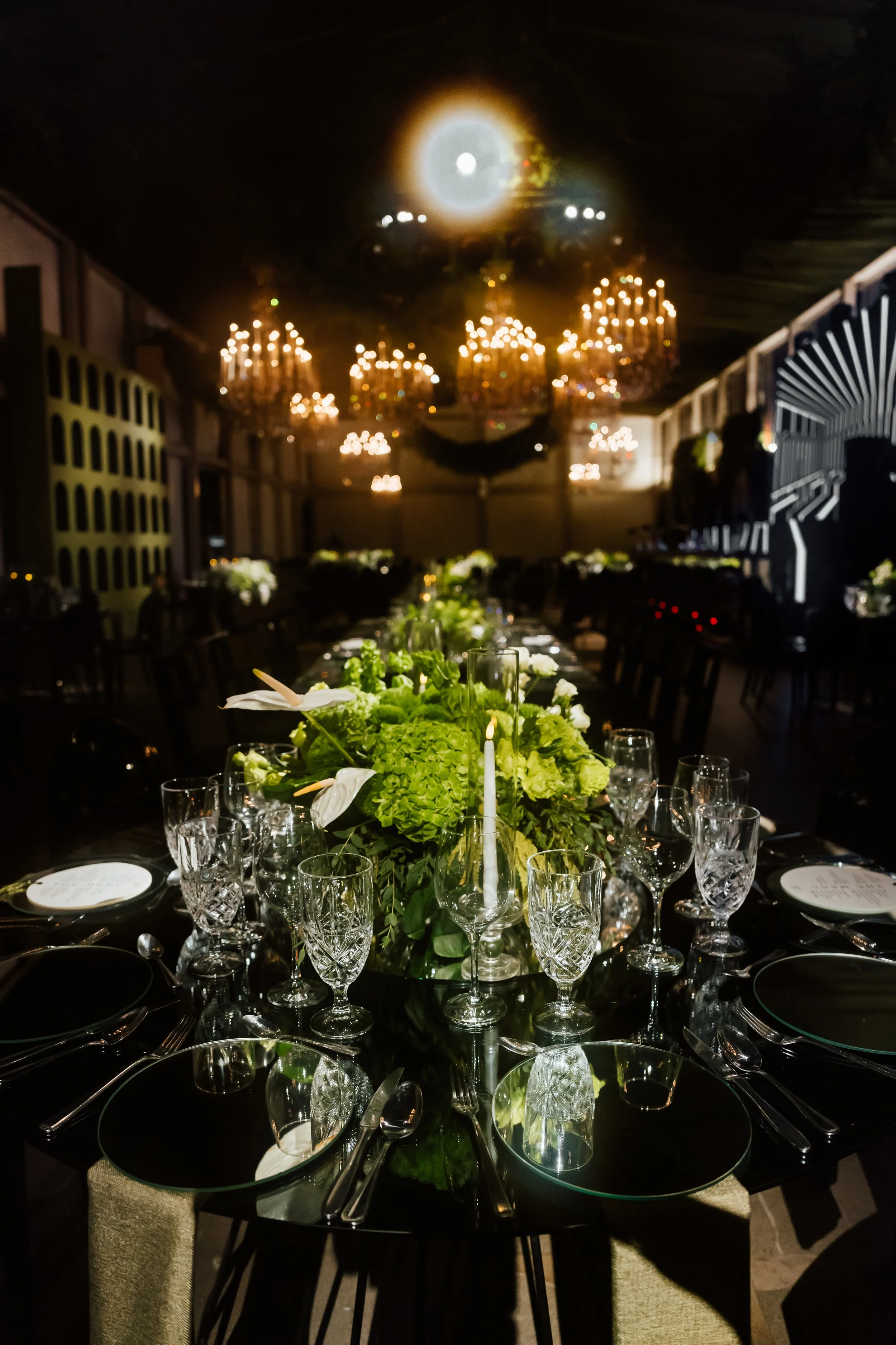 Elegant banquet table set with green floral centerpiece, crystal glassware, plates, and silverware, under chandeliers in a dimly lit room.