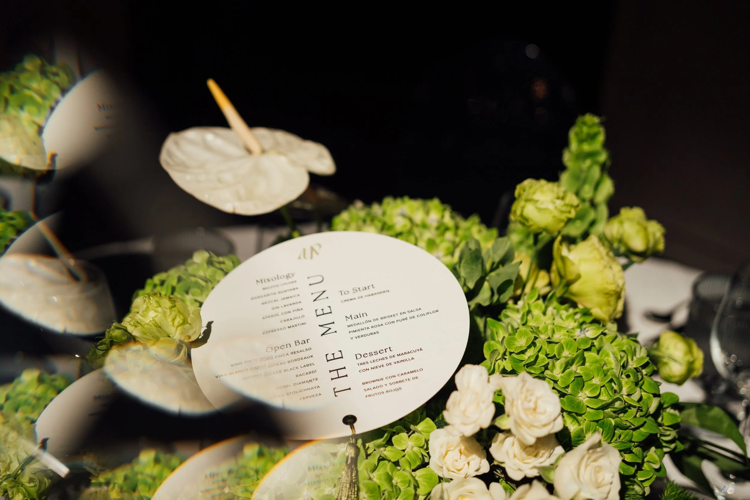 A floral arrangement includes cream-colored roses and green hydrangeas surrounding a round menu card on a black surface.