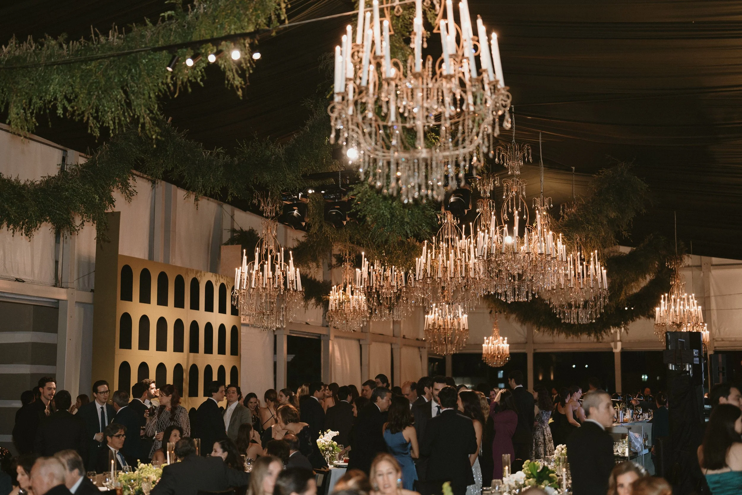 Elegant indoor event with large crystal chandeliers hanging from a dark ceiling, surrounded by greenery. Many people in formal attire socialize at tables with floral centerpieces.
