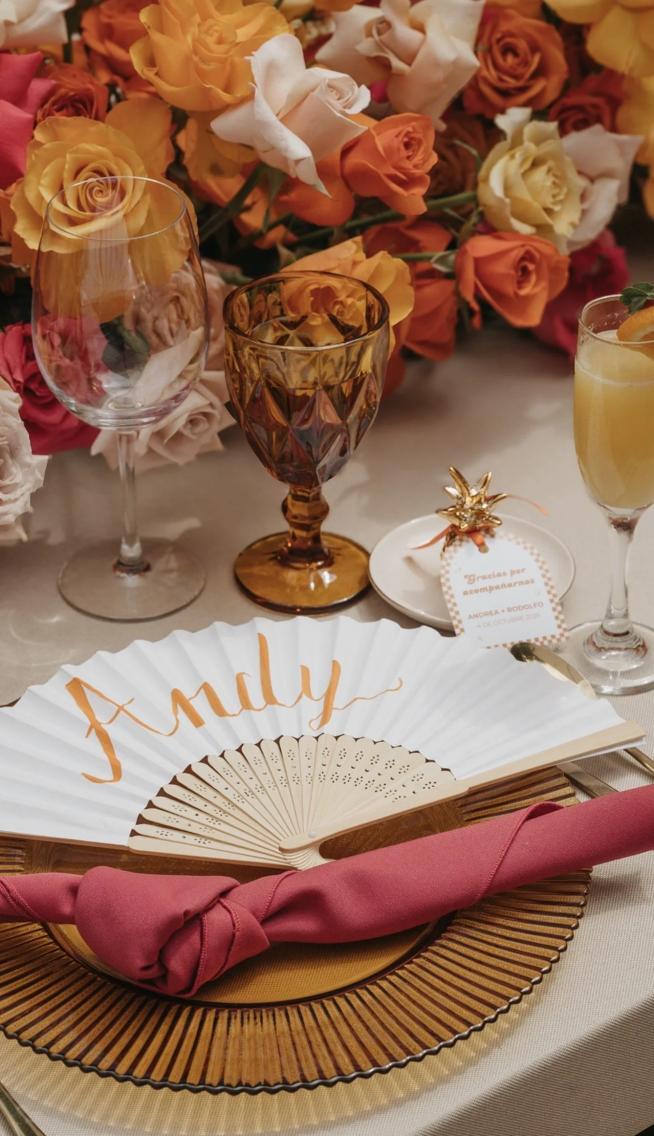 A table setting with colorful roses, wine glasses, a drink, a decorative fan with the name 'Anully,' a pink cloth napkin, and an assortment of place cards and decorations for a celebration.