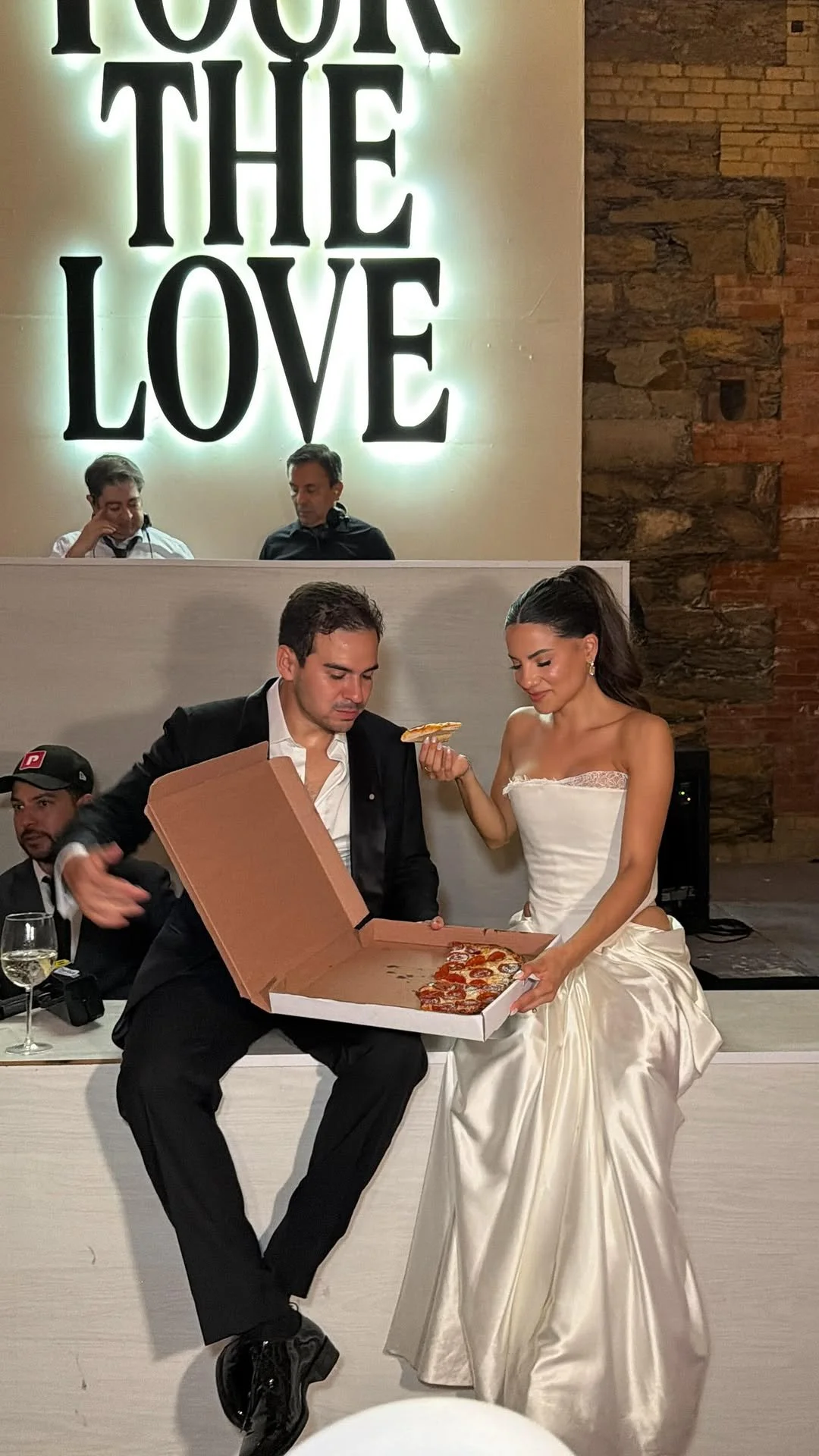 A bride and groom in wedding attire sharing a pizza at their wedding reception, with the bride smiling and holding a slice of pizza, and the groom opening a pizza box.