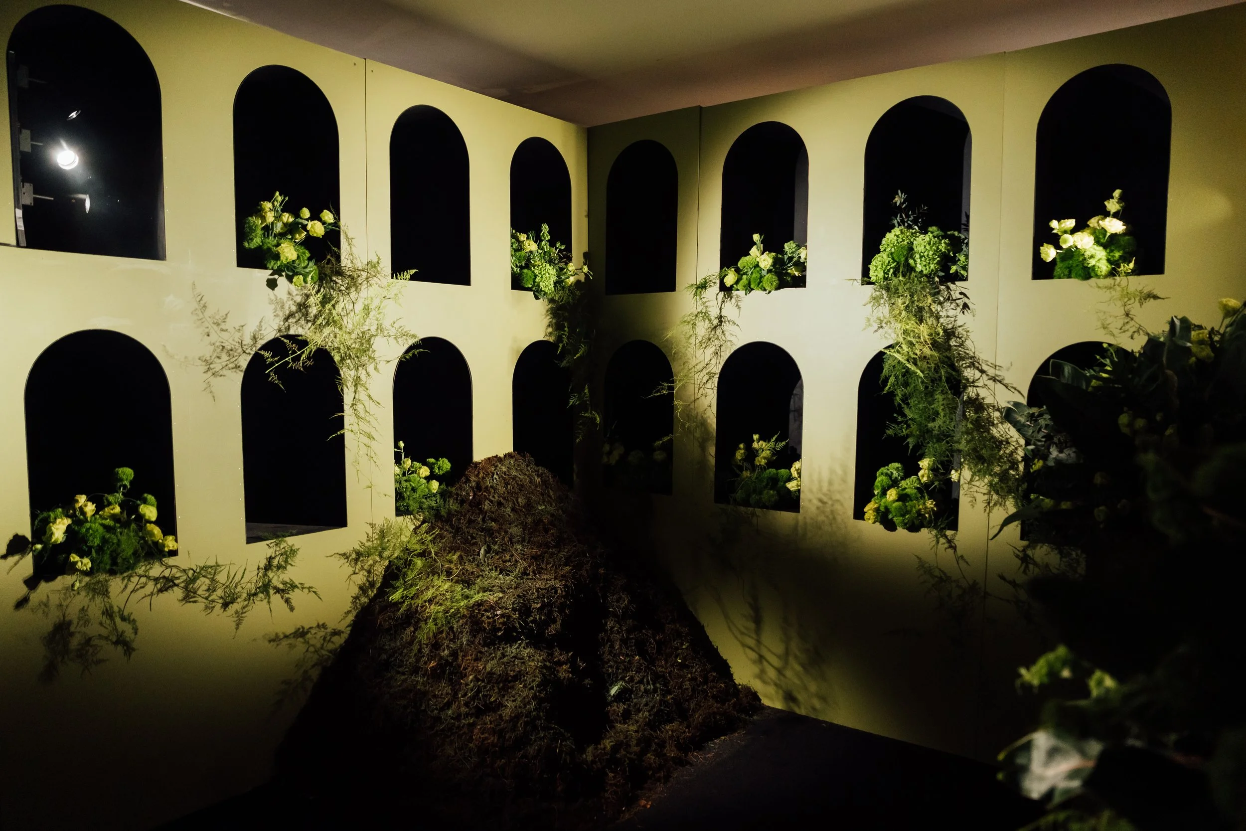 An art installation featuring a wall with multiple arched openings filled with green plants and flowers, illuminated by focused lighting, with a mound of soil and plants in the foreground.