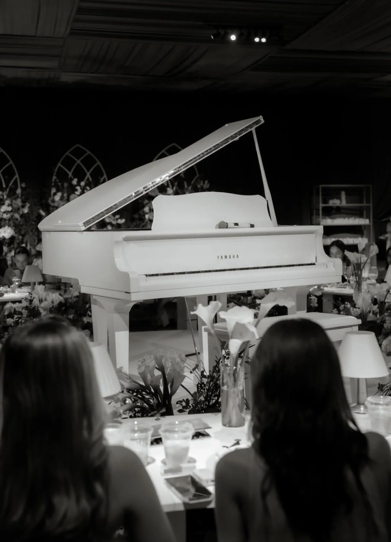 A white grand piano on a stage with two women sitting at a table in the foreground. The stage is decorated with flowers and lamps, and there are people watching the performance.