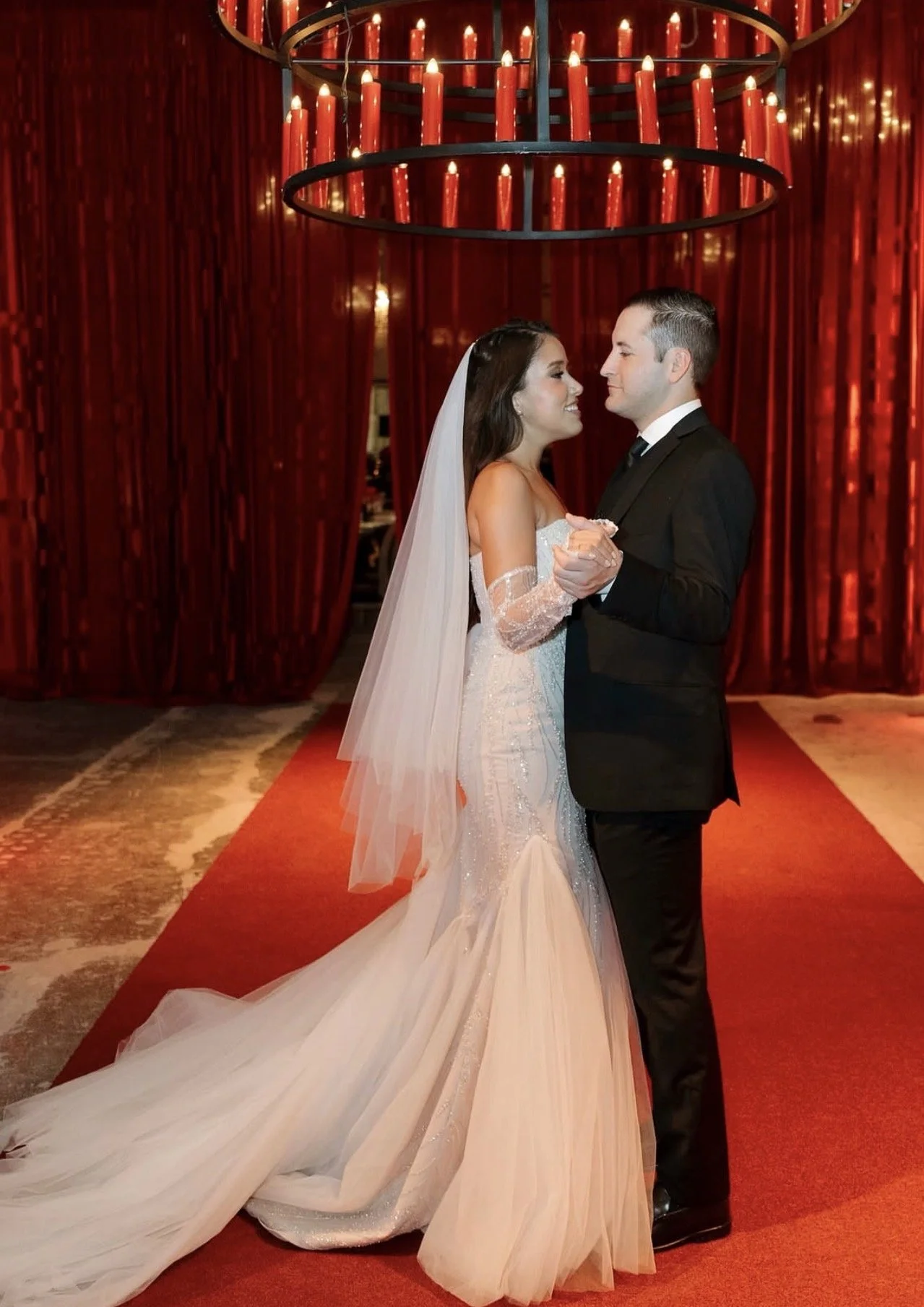 Bride and groom dancing at their wedding reception, standing on a red carpet beneath a large circular chandelier with red candles, with red curtains in the background.