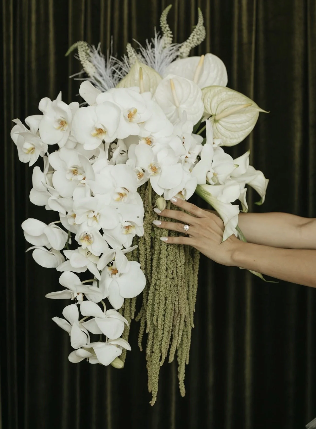 A bouquet of white orchids, anthuriums, and other white flowers being held by a person with a wedding ring, set against a dark background.