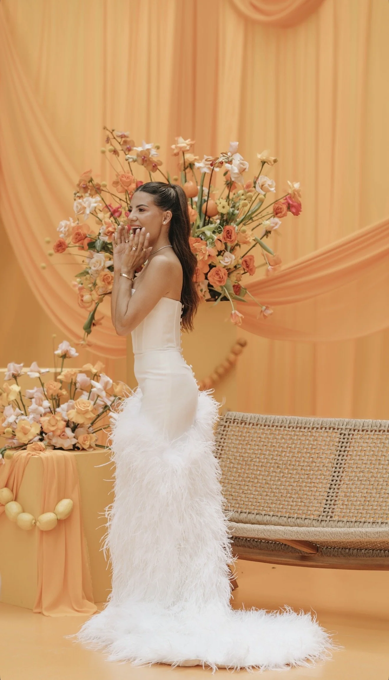 Woman in a white strapless dress with feathered train, standing in front of a peach-colored draped background and floral arrangement, smiling and covering her mouth with her hands.
