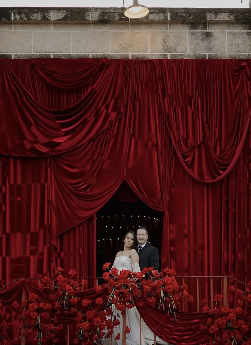 A bride and groom on a wedding balcony surrounded by red roses and draped in red fabric, smiling and sharing a tender moment.