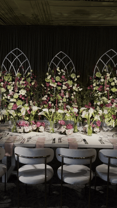 Elegant banquet table decorated with pink and white floral arrangements, calla lilies, and greenery, set against a black backdrop with white lattice arches.