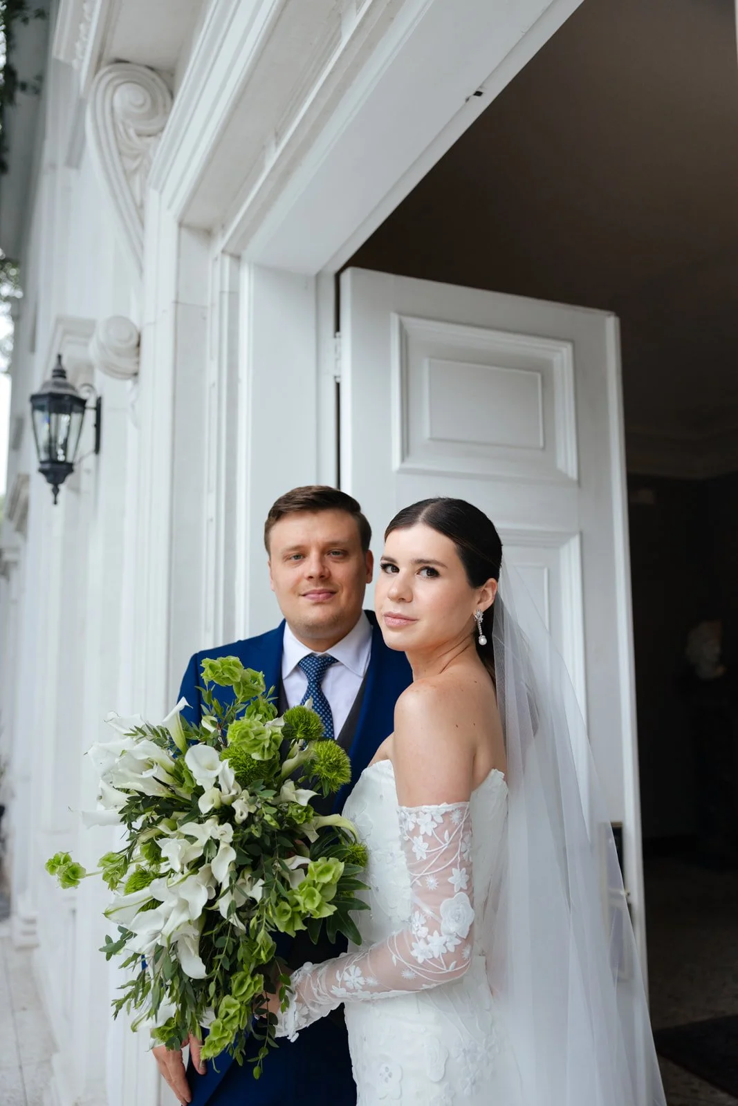A bride and groom standing together in wedding attire, outside a building with white trim. The bride is holding a large bouquet of white and green flowers, wearing off-the-shoulder lace wedding gown and veil. The groom is wearing a dark blue suit wit