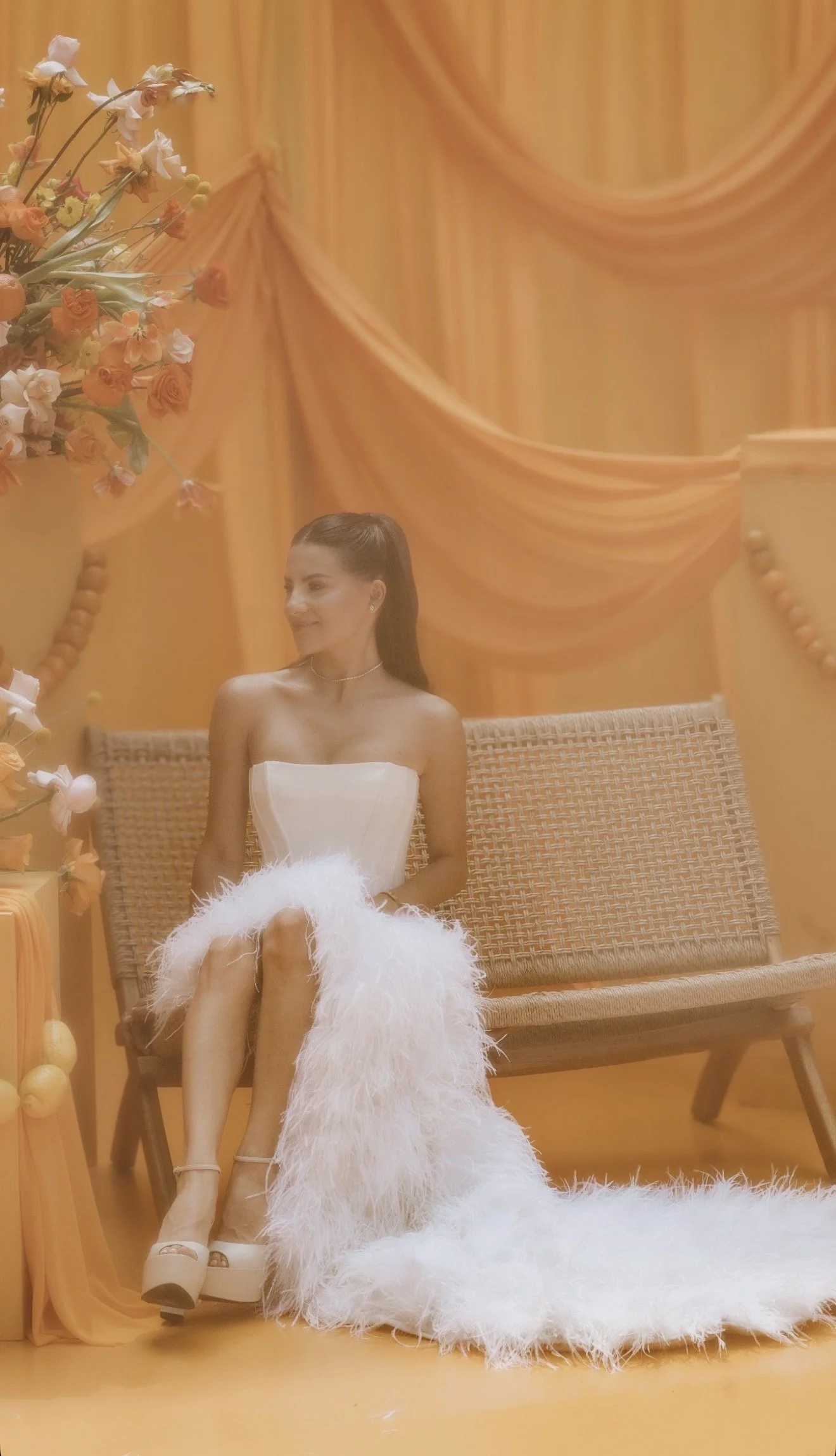 A young woman in a white strapless dress with a fluffy hem, sitting on a wicker bench in a room with orange curtains and floral decorations.