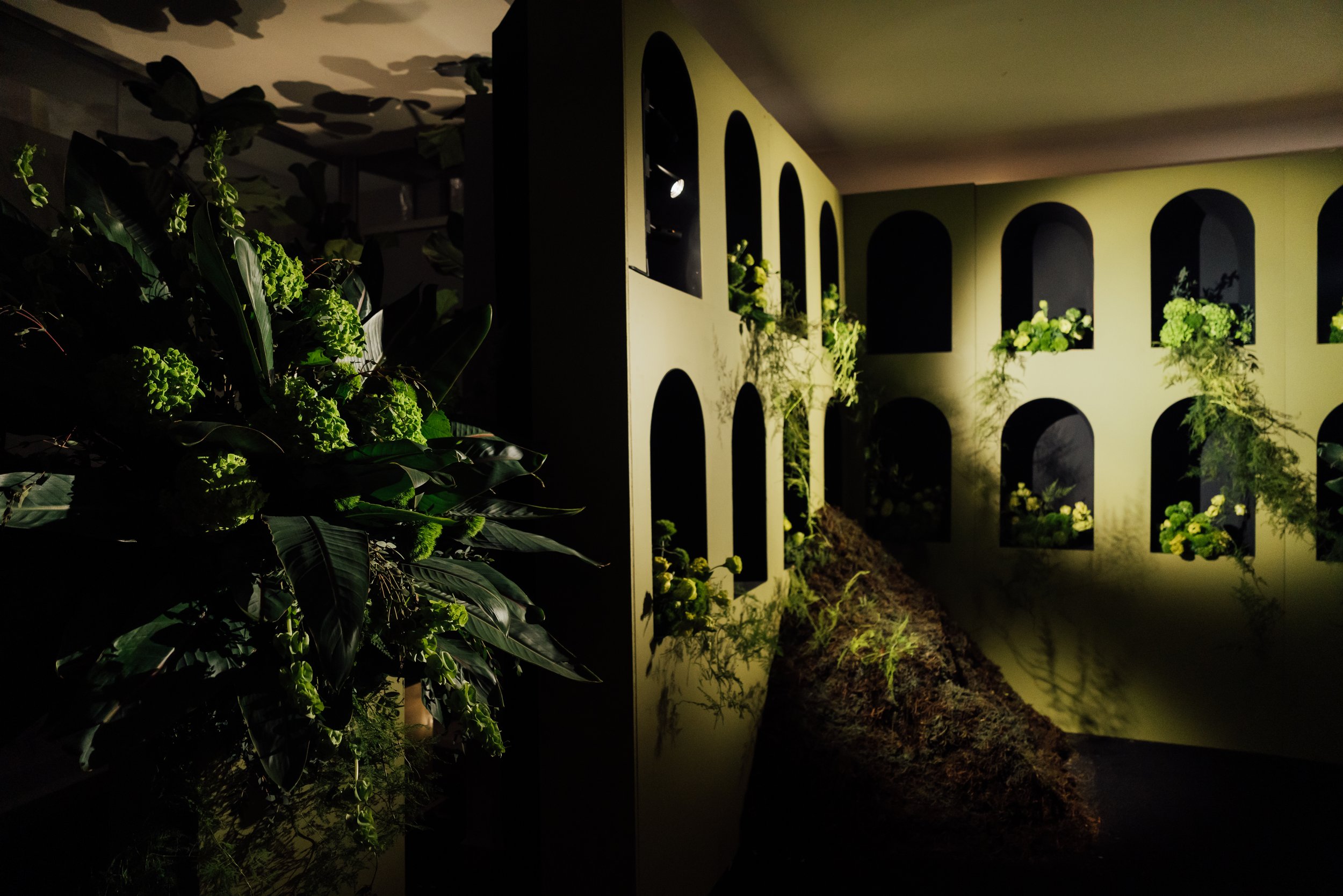 Indoor display with botanical arrangements and a structure resembling a small building with arched windows, illuminated by soft lighting.