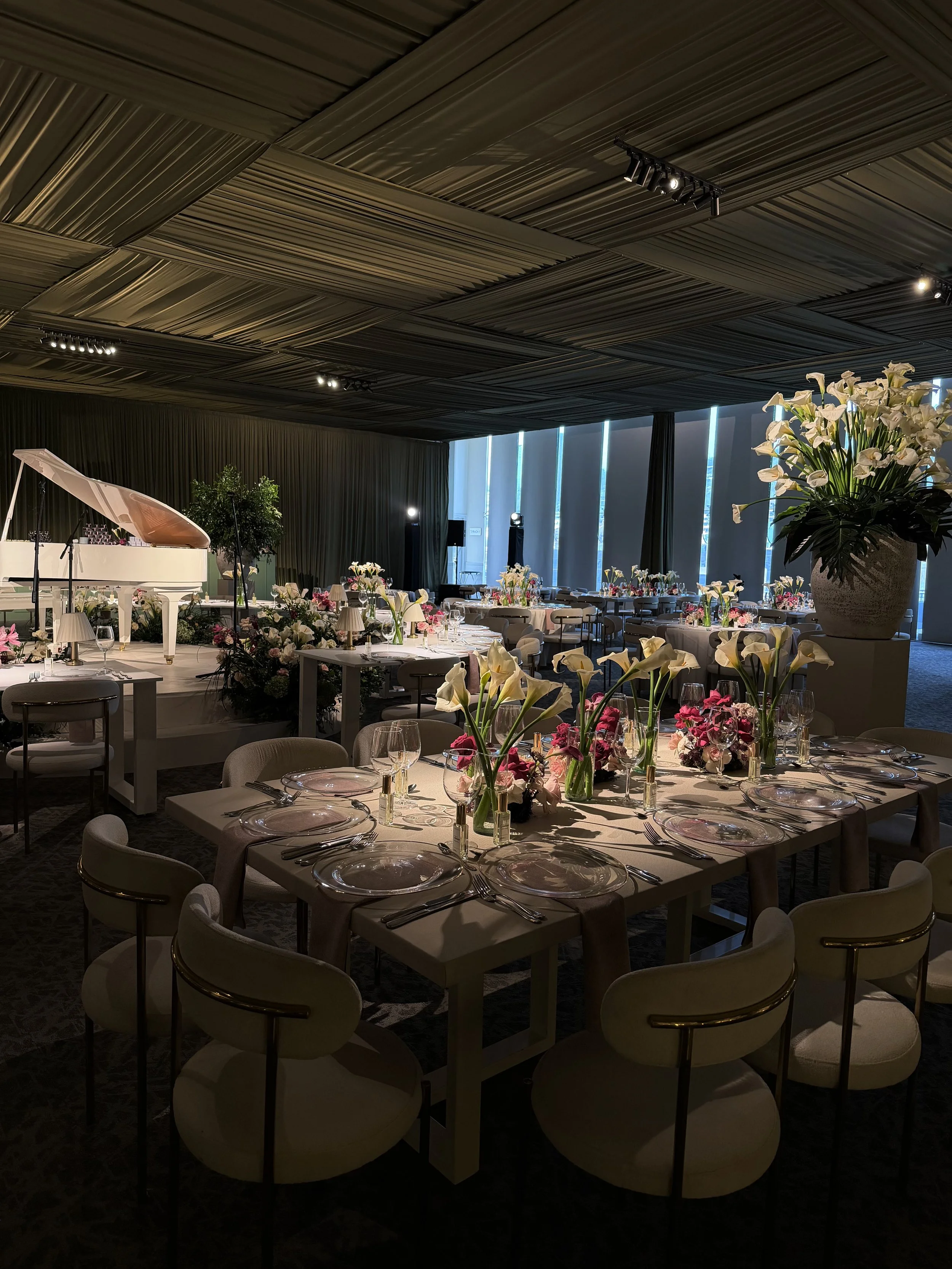 Indoor event space decorated with floral arrangements, featuring tables set with glassware and plates, and a white grand piano on a stage with black curtains and vertical window panels in the background.