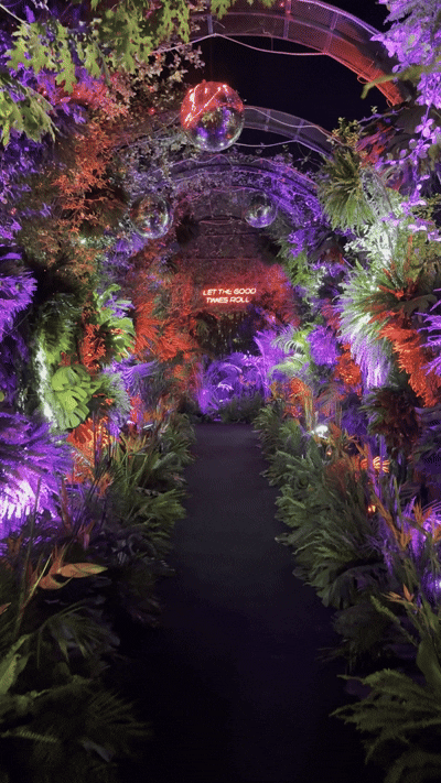 A vibrant botanical tunnel adorned with purple, pink, and red foliage, colorful lights, hanging disco balls, and a neon sign that reads 'Let the good times roll'.