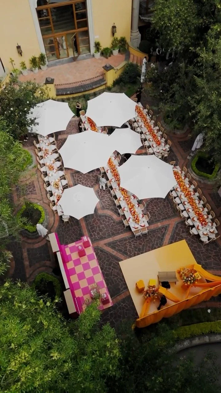 Outdoor event setup with large white umbrellas covering round tables decorated with orange and yellow flowers. A pink checkered platform is on the left, and a yellow stage with floral arrangements is at the bottom. The area is surrounded by trees and potted plants.