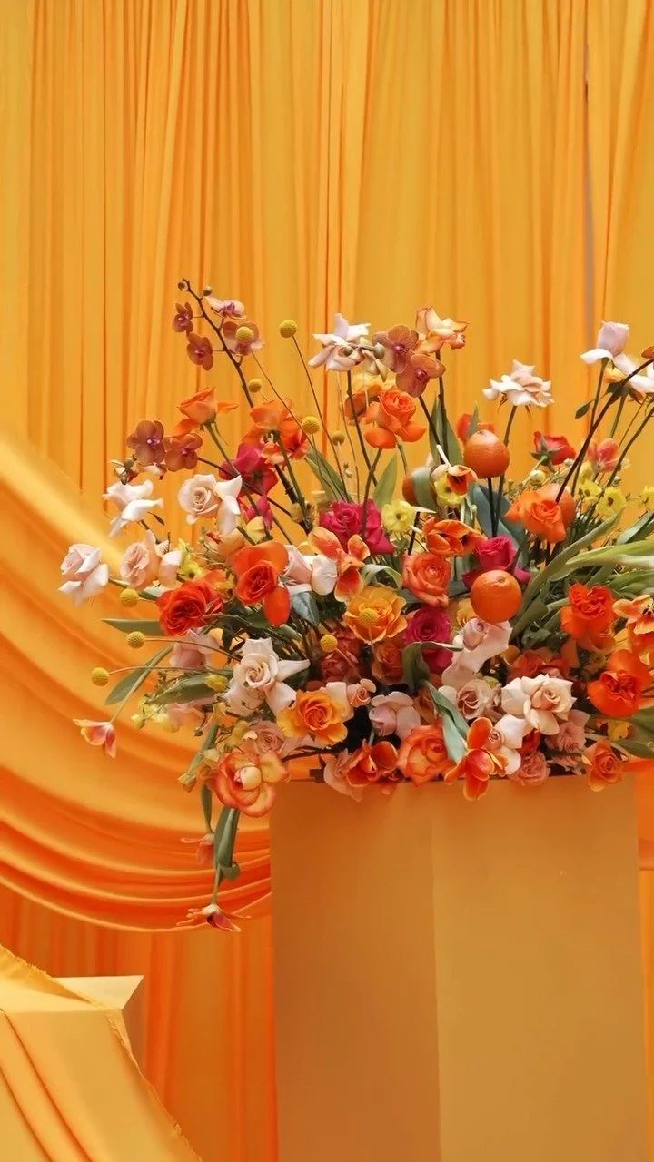 A bright yellow floral arrangement with roses, orchids, small oranges, and greenery in a tall yellow vase, set against a yellow draped background.