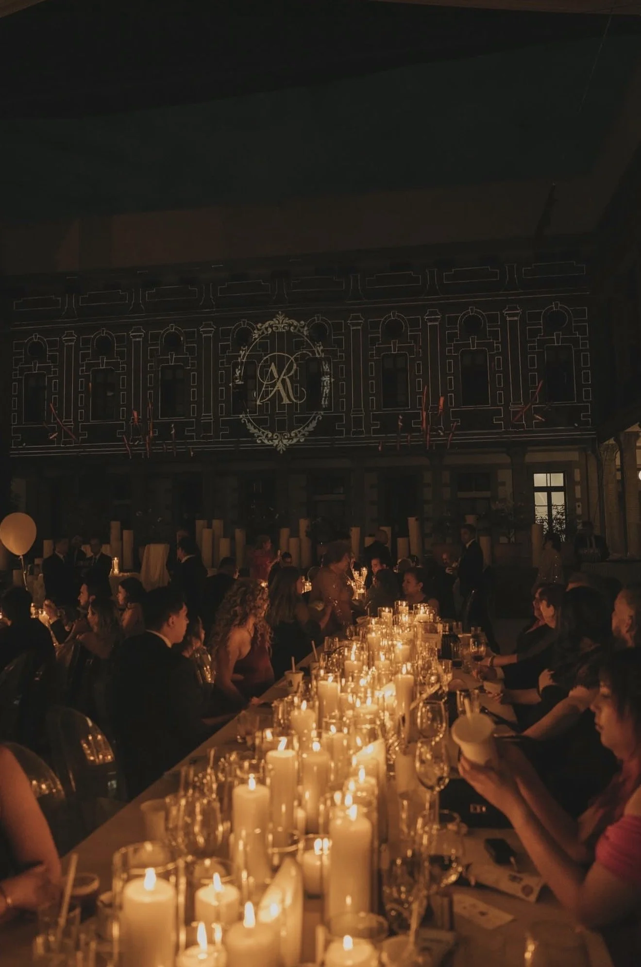 People seated at a long table with numerous lit candles, dining at a formal event in a dimly lit room with candles and soft lighting.