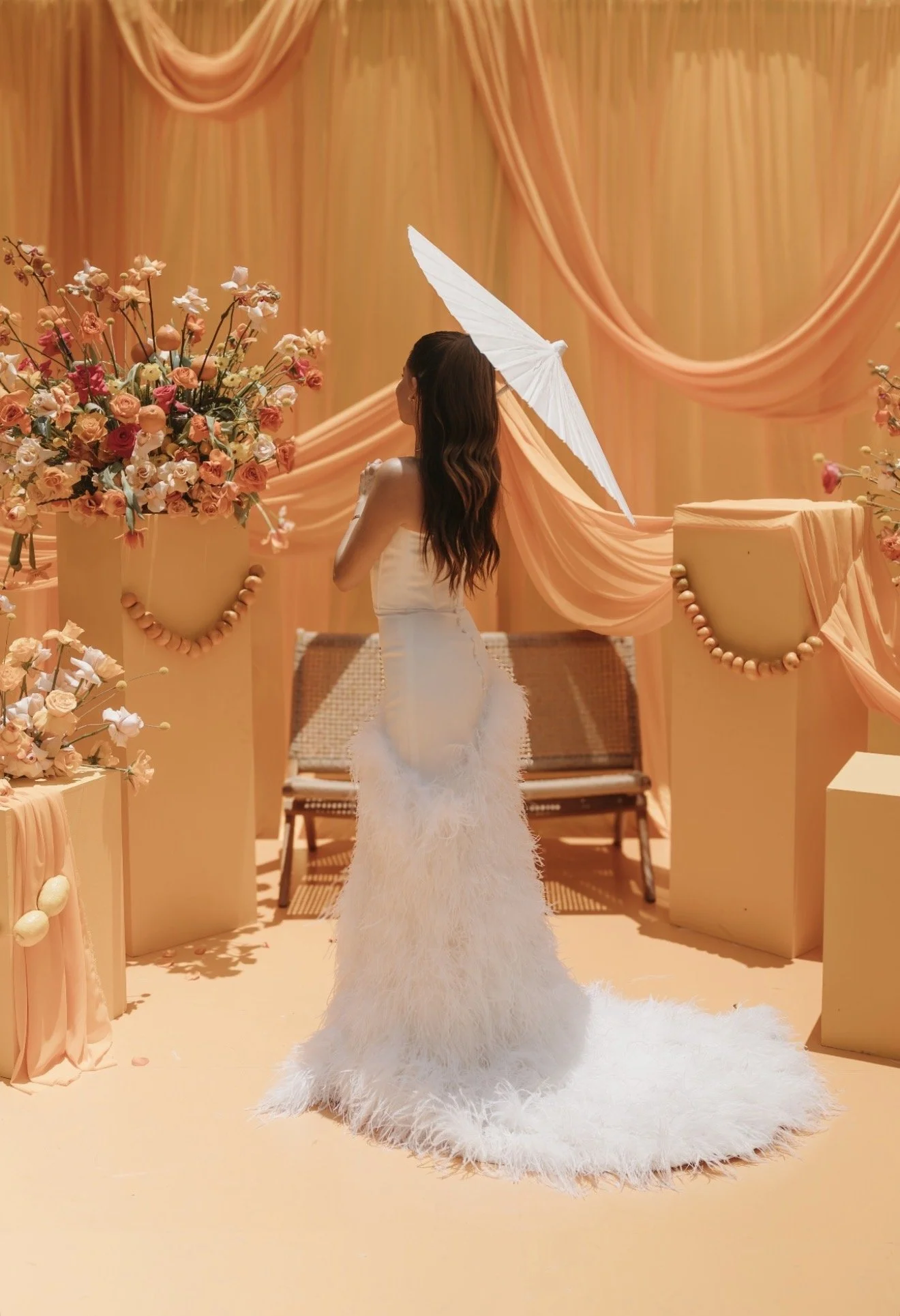 A woman in a white feathered gown holding a white parasol standing in front of peach-colored drapes with floral arrangements on either side.