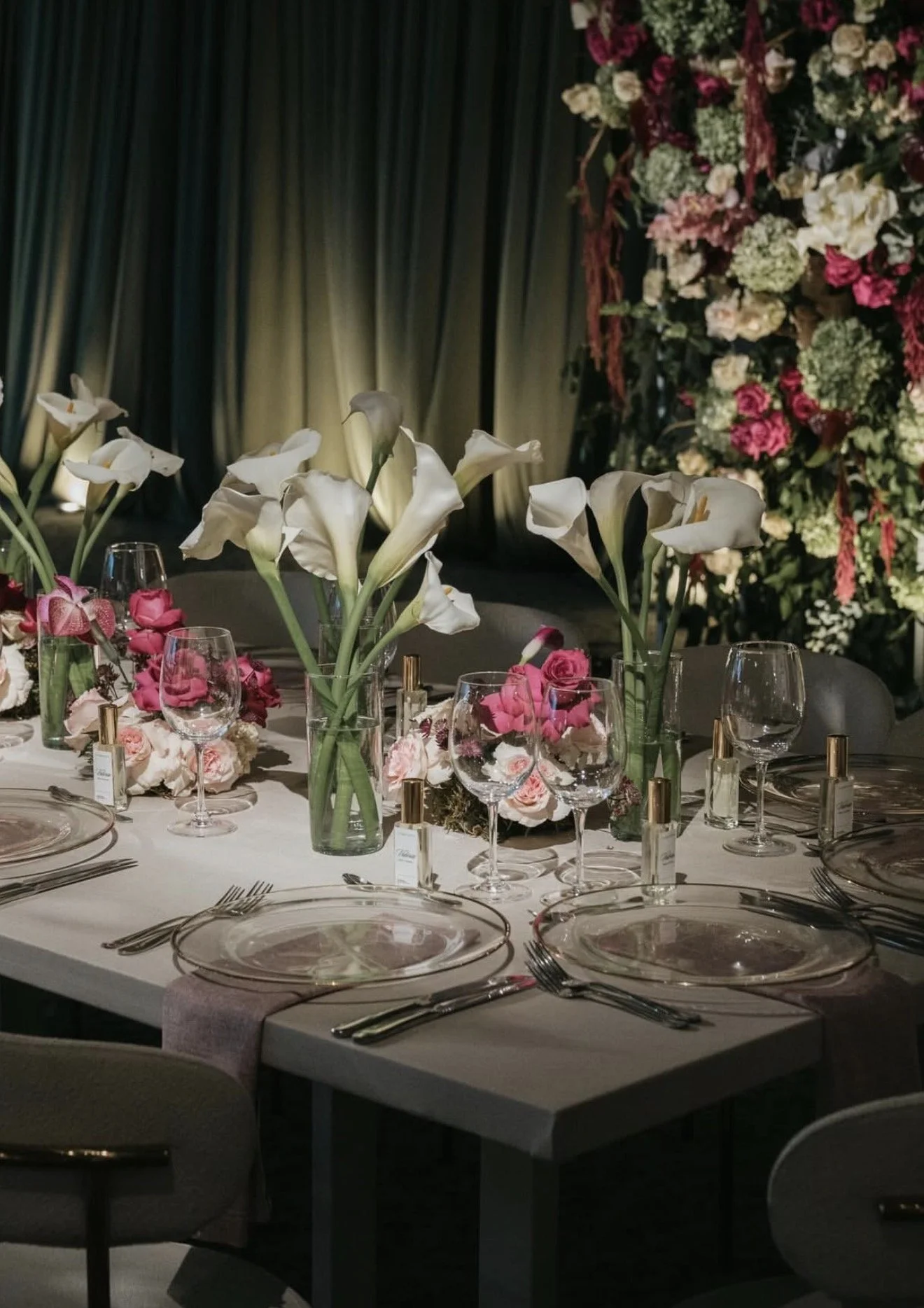 Elegant banquet table with white tablecloth, floral centerpieces of white calla lilies and pink roses, surrounded by glassware, plates, and utensils, with a background of draped curtains and an arrangement of mixed flowers.