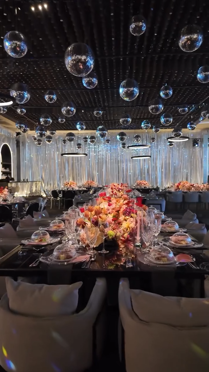 A decorated dining table with floral centerpieces, set with glassware and plates, in a dimly lit room with silver curtains and colorful light reflections.