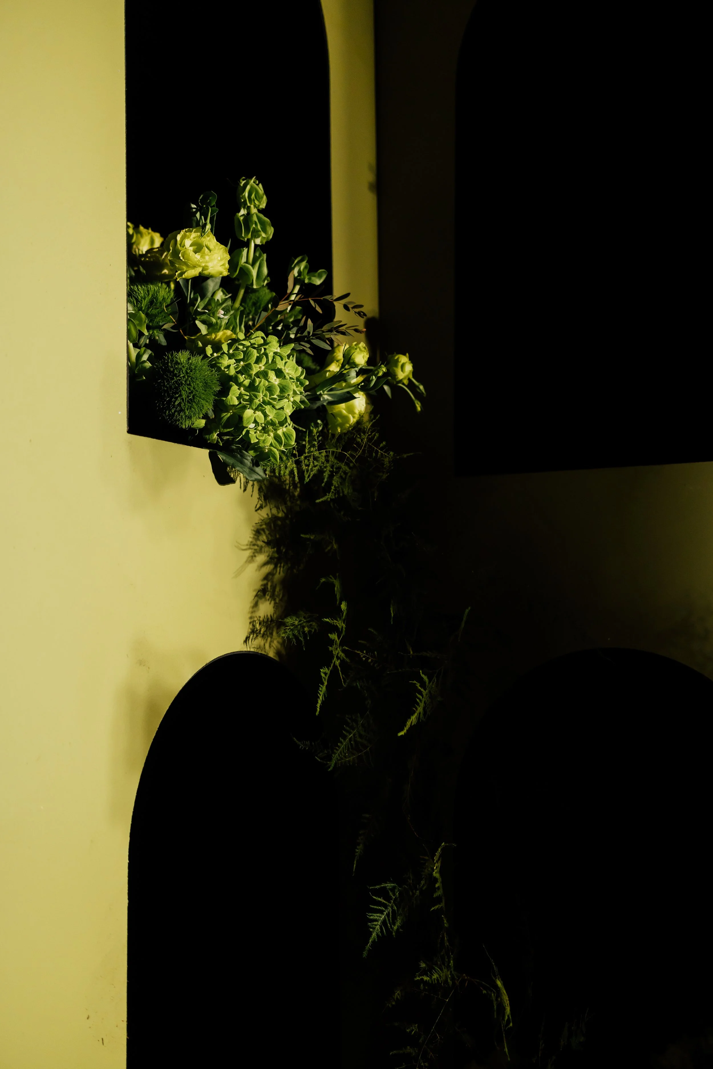 A floral arrangement with white and green flowers and foliage in a black rectangular container, placed on a light-colored surface with shadows cast on it.