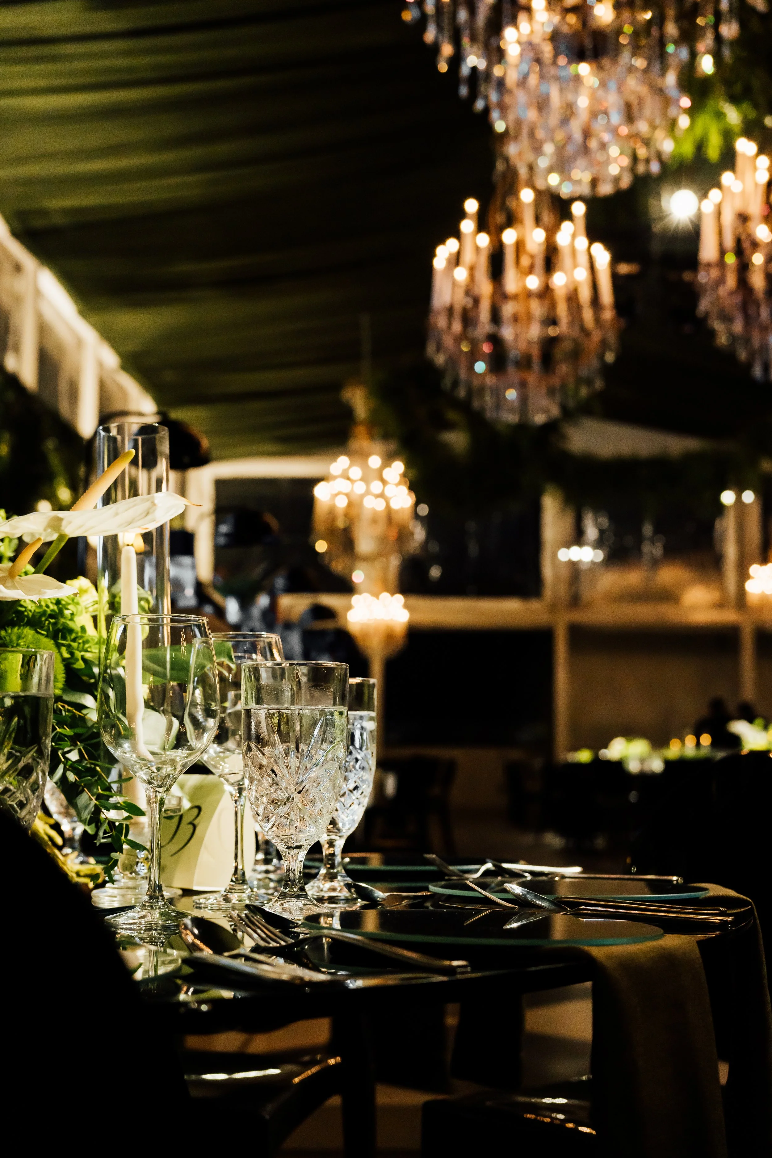 Elegant table set with wine glasses, silverware, and floral arrangements in a dimly lit room with chandeliers hanging from the ceiling.