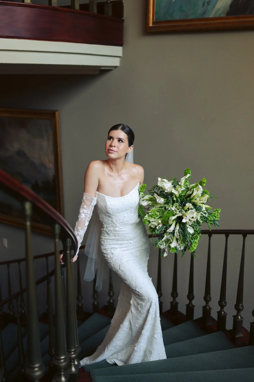 A woman in a wedding dress standing on a staircase with a large bouquet of white and green flowers.
