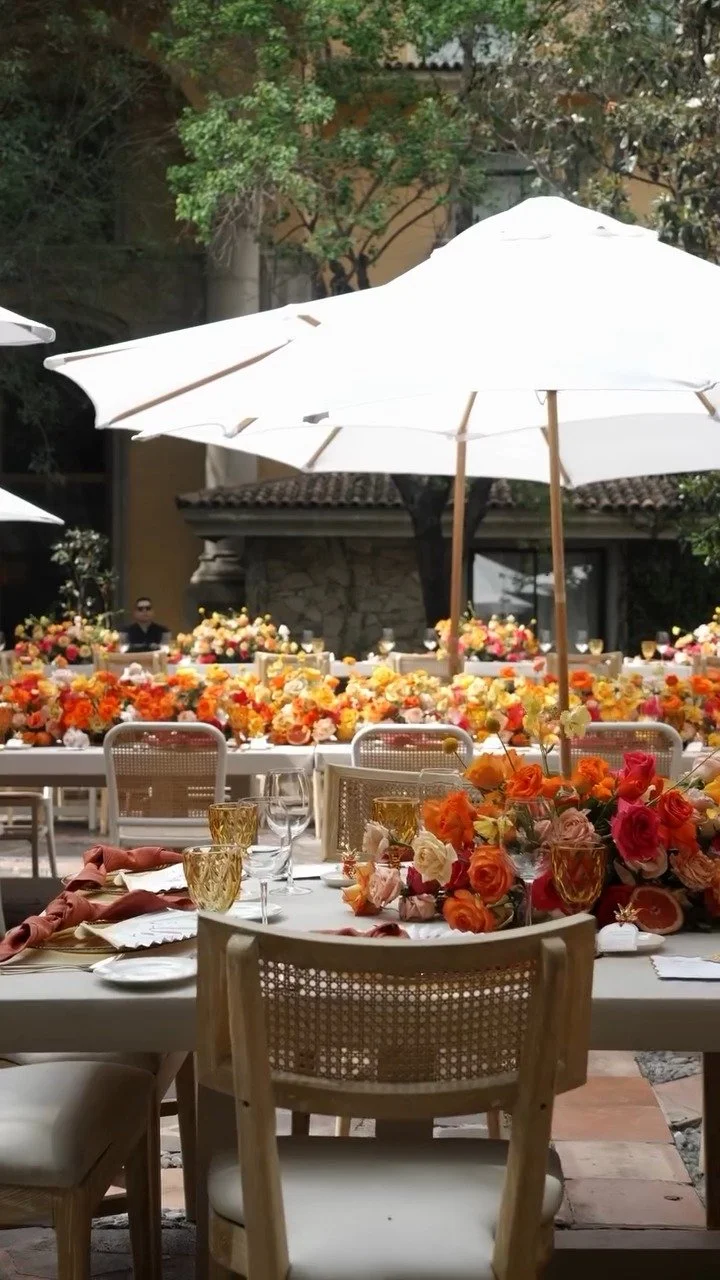 Outdoor event setup with decorated tables, floral centerpieces, glasses, and large white umbrellas during daytime.