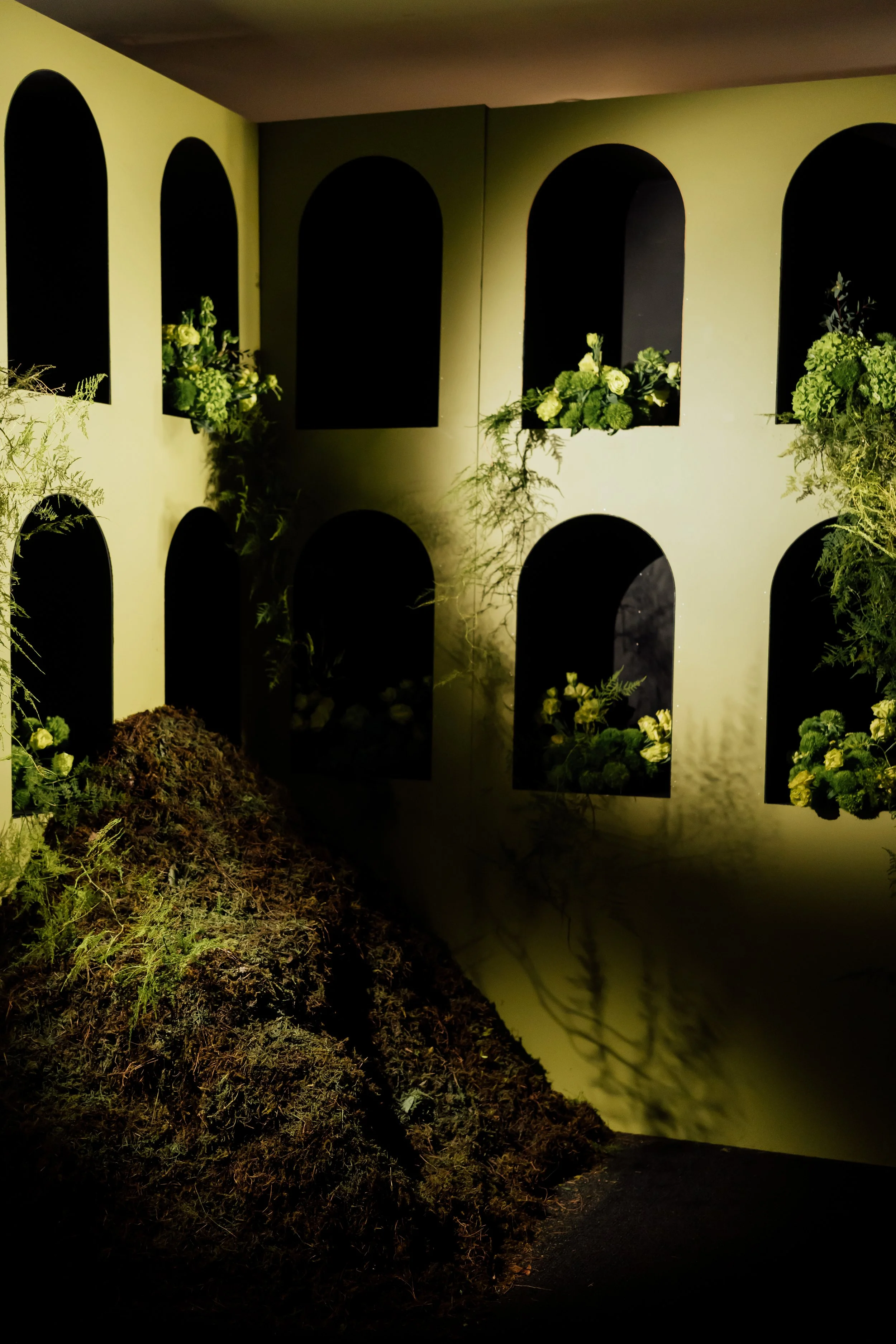 Display of small niches with floral arrangements in a wall, with some plants and moss on the ground in front.