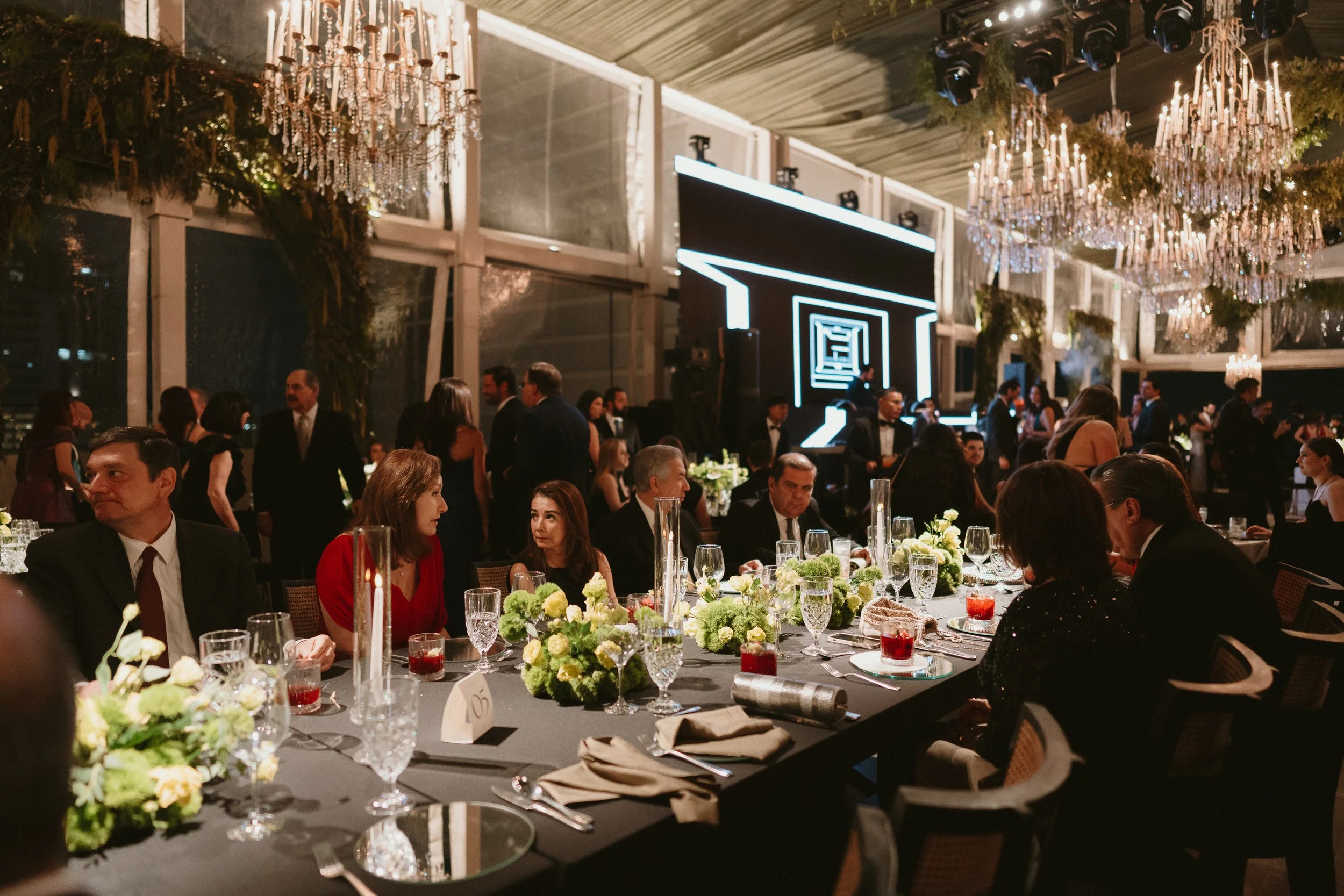 A formal dinner event at a banquet hall with elegant chandeliers, a long table set with floral arrangements, glasses, and dinnerware, and guests dressed in formal attire engaging in conversation.