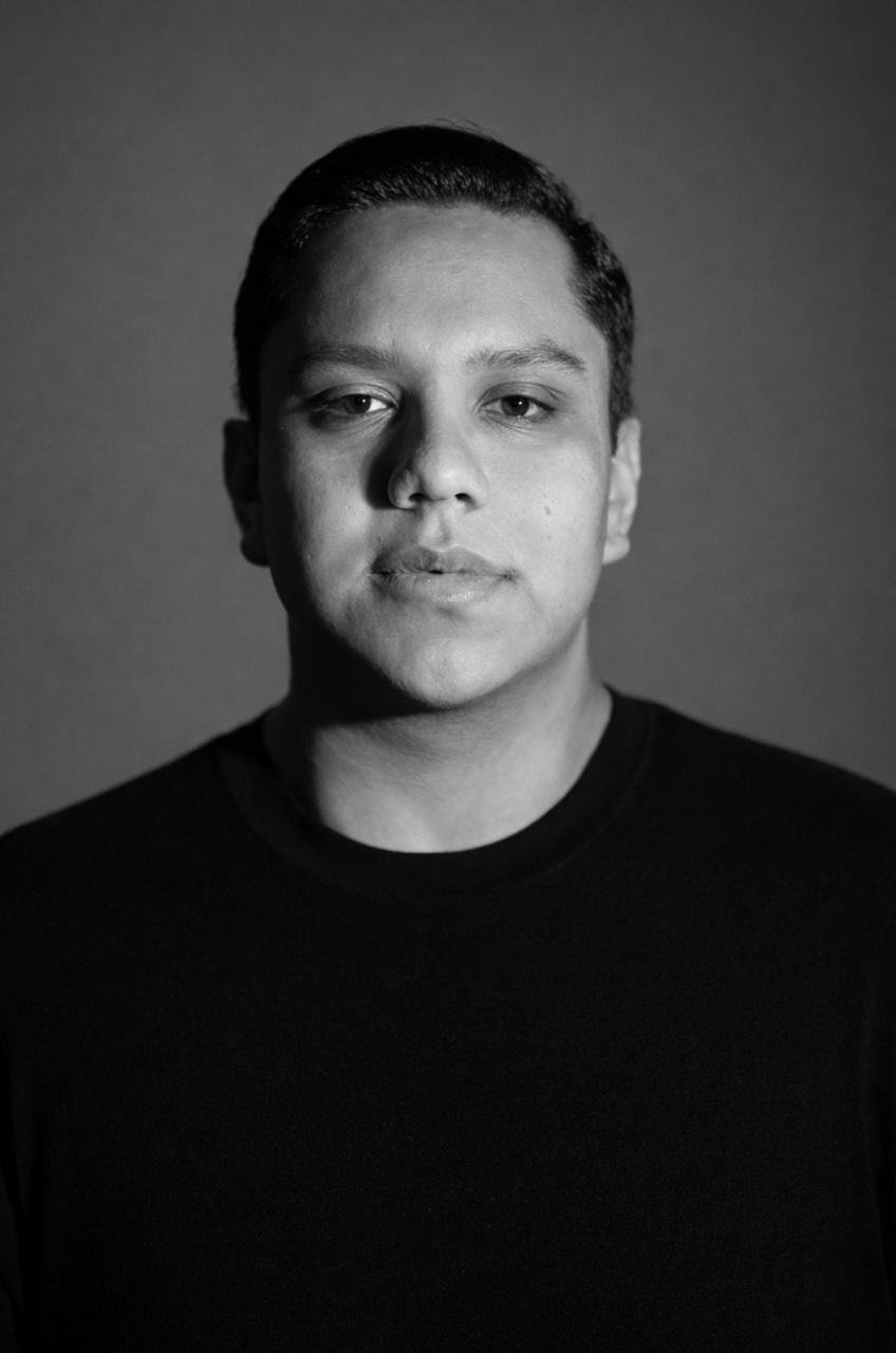 Black and white portrait of a young man with short dark hair, wearing a black shirt, looking directly at the camera with a neutral expression.