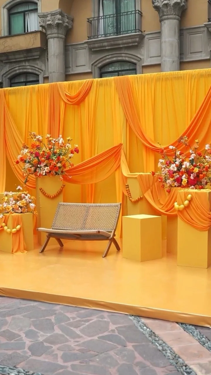Decorated stage with orange drapes, flower arrangements, and a small bench, set against an urban building backdrop.