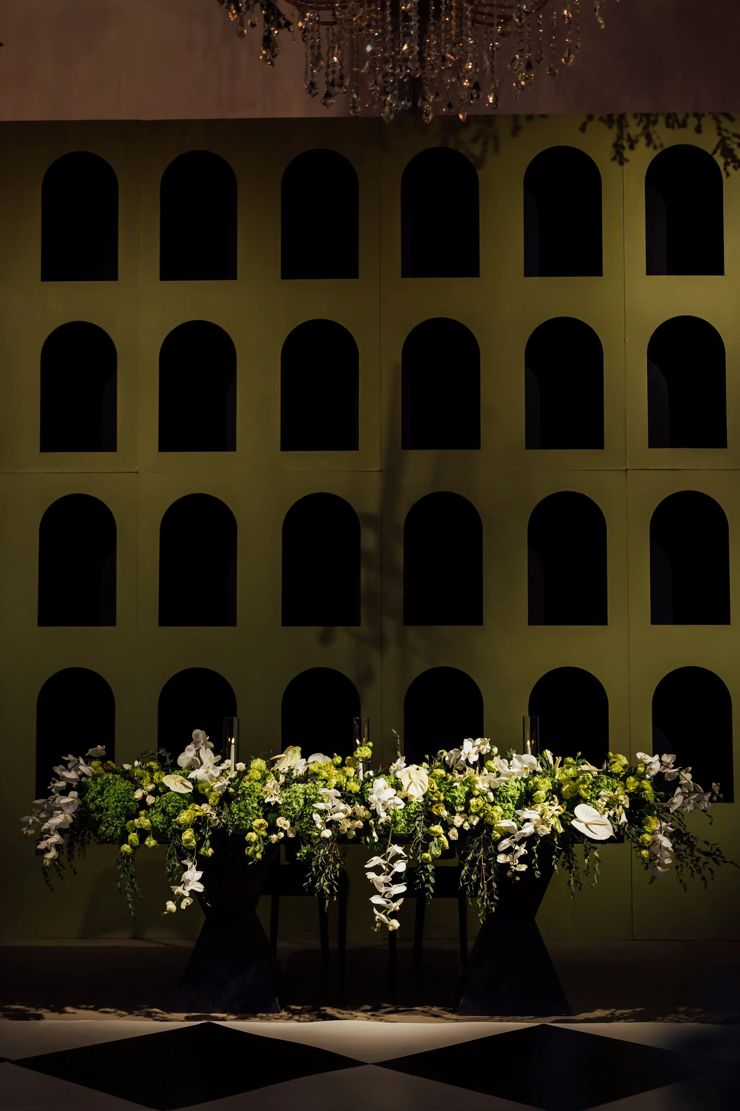 Elegant floral arrangement of white and green flowers and foliage in black vases, placed in front of a wall with a grid of black arched openings, with a chandelier hanging above.