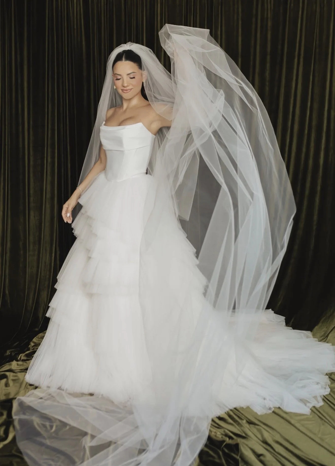 A smiling woman in a white wedding dress and veil standing in front of gold and black curtains.