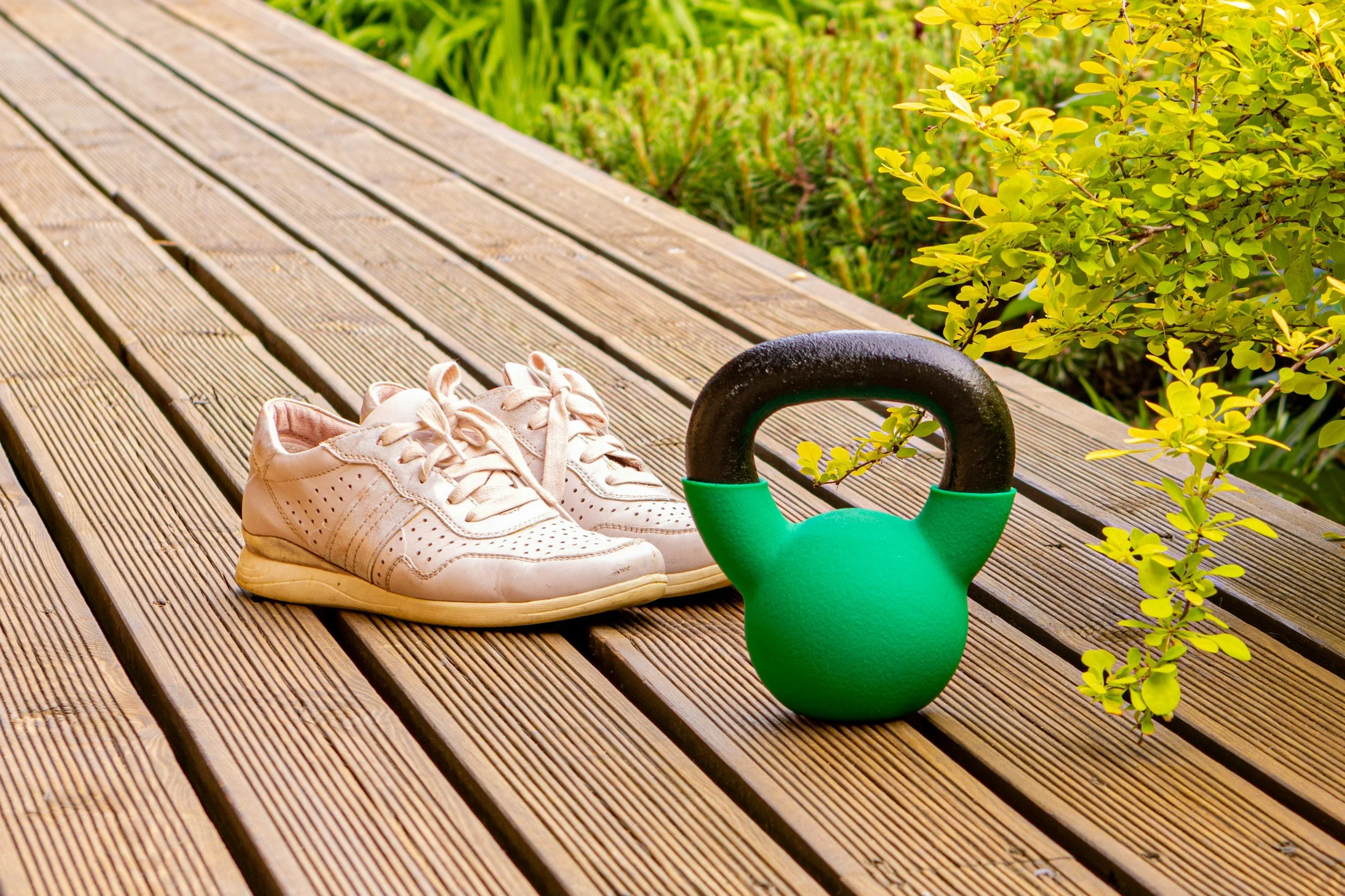White athletic shoes, a green kettlebell, and yellow-green foliage on a wooden deck.