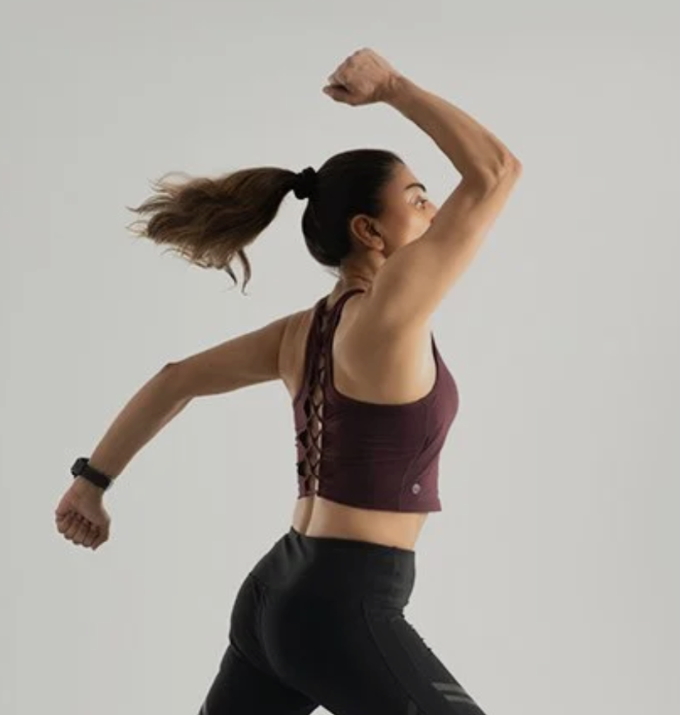 A woman running outdoors, wearing a sports bra and black athletic shorts, with her hair in a ponytail and jumping with her arms raised.