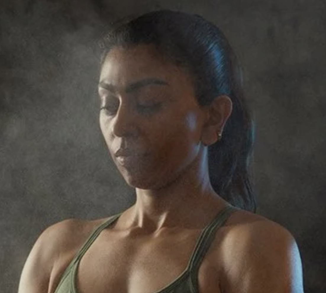 A woman with dark hair in a high ponytail, wearing a green tank top, with her eyes closed, against a dark background.
