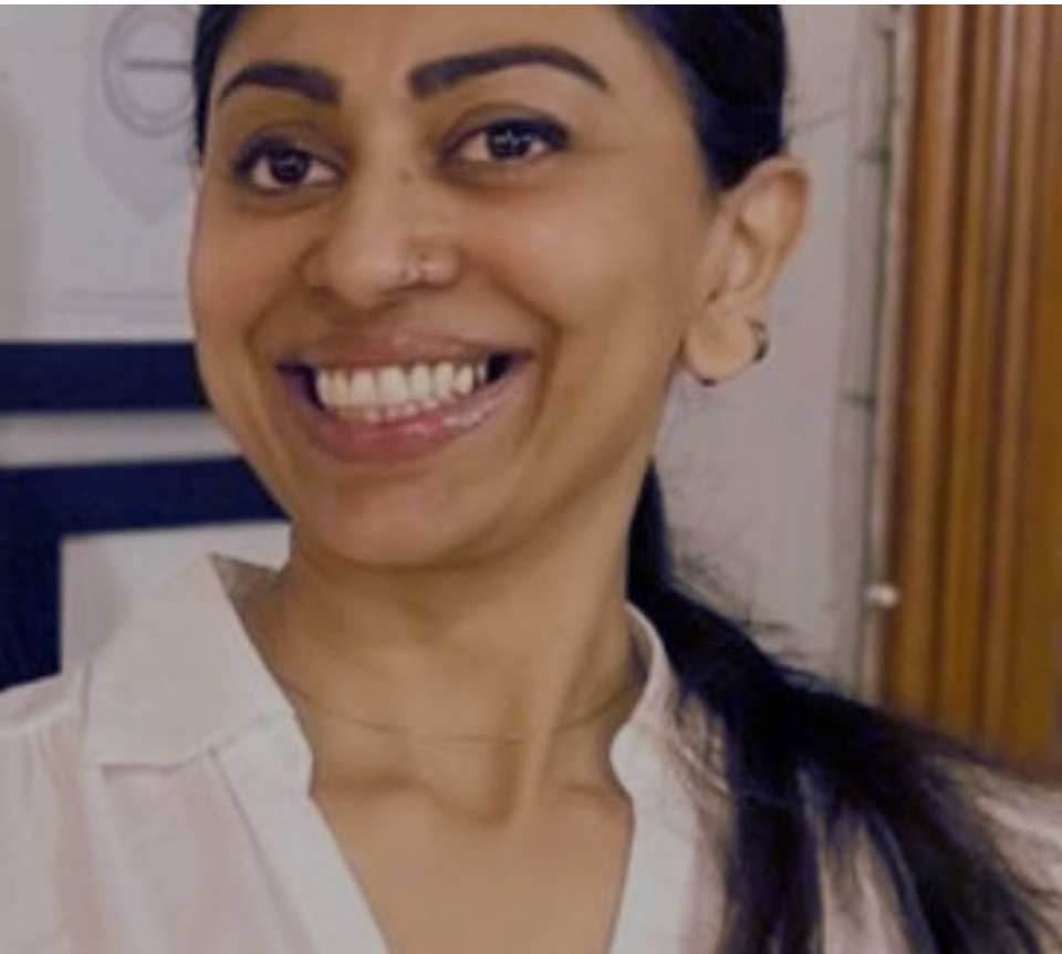 A woman smiling with dark hair pulled back, wearing a white top, standing indoors near a wooden door and a poster on the wall.