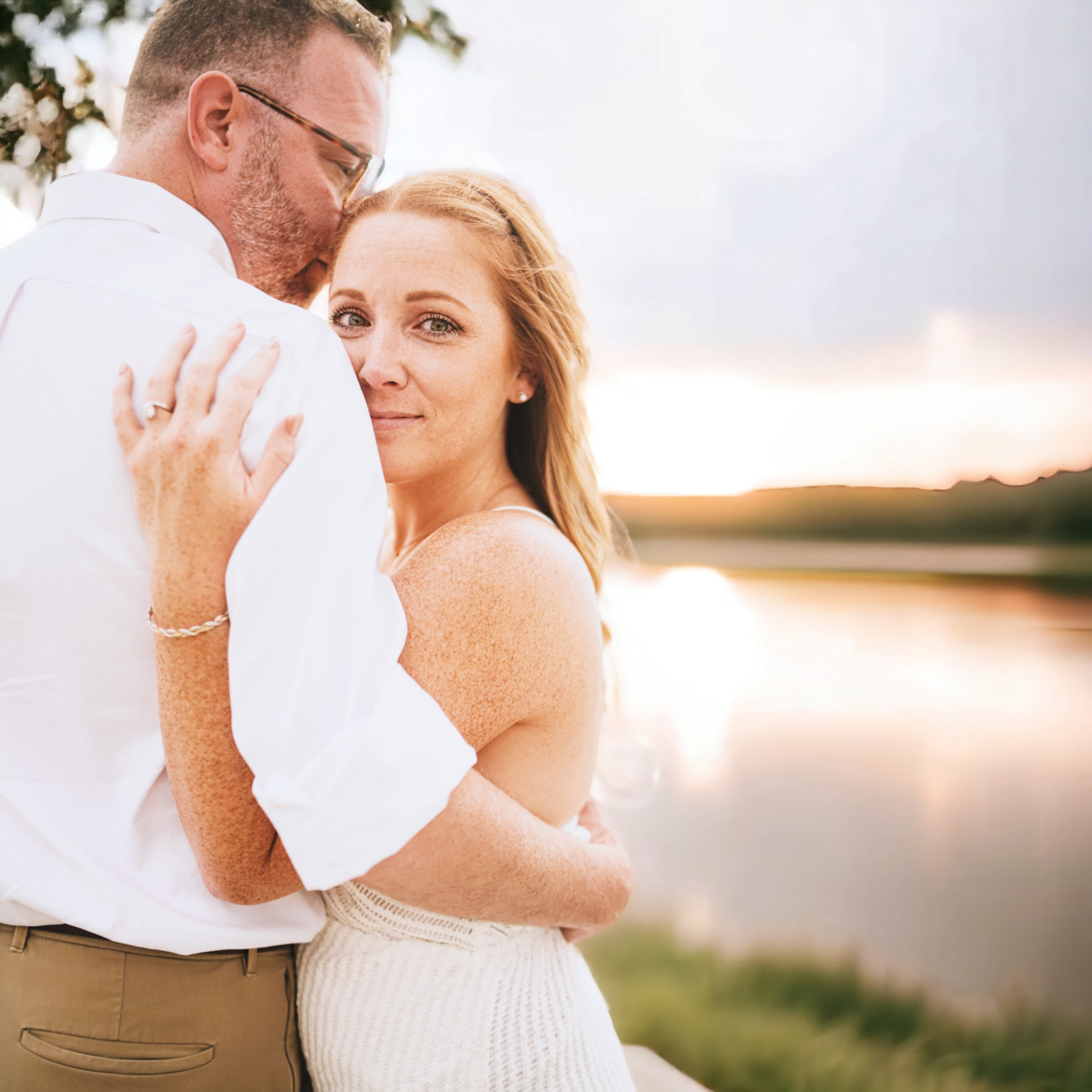 A couple embracing outdoors during sunset by a body of water, with lush green landscape in the background.