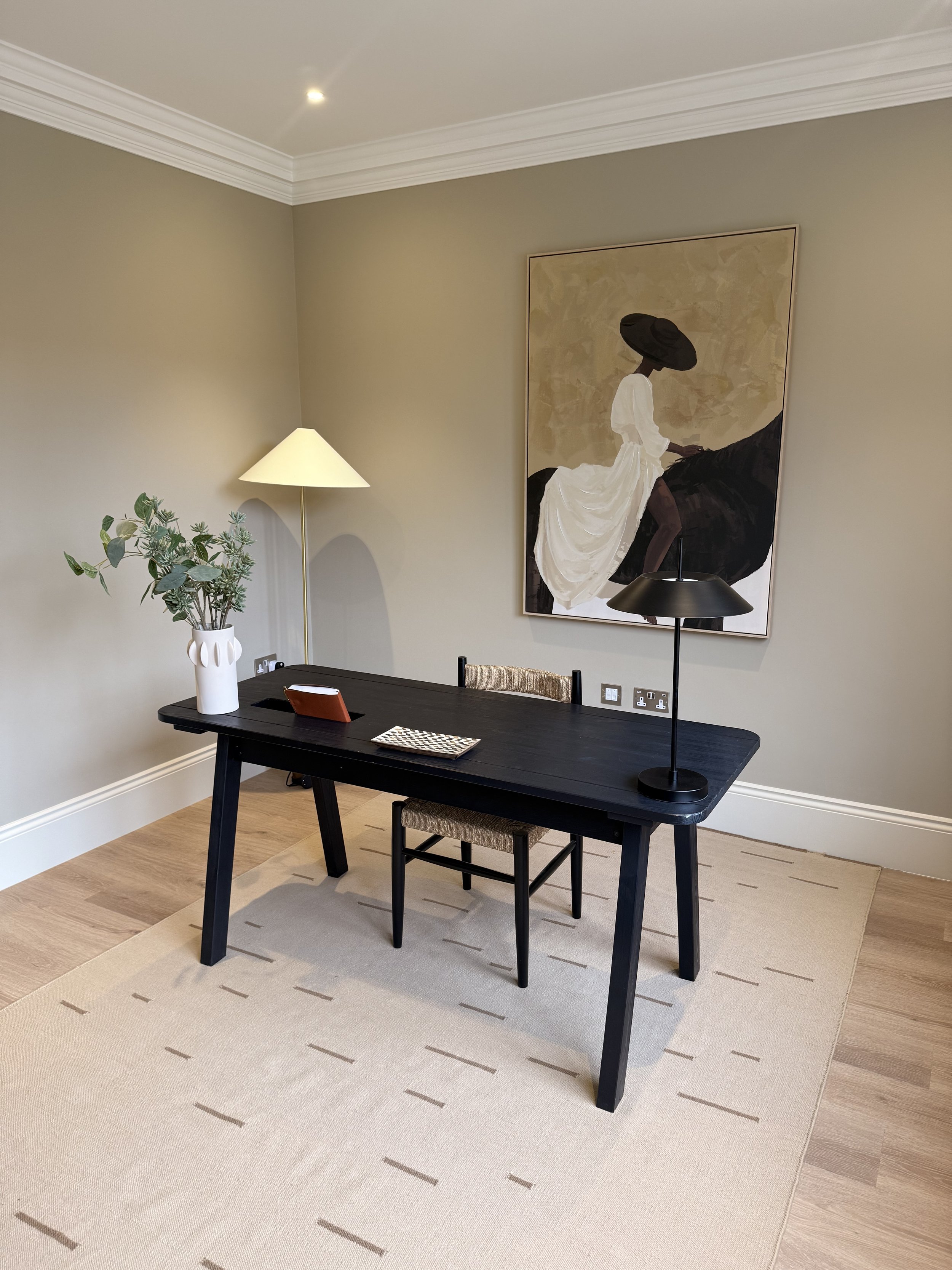 A dining room with a black table, beige area rug, and a painting of a woman in white dress and black hat mounted on a beige wall. A floor lamp with a white lampshade and a potted plant are on the table.