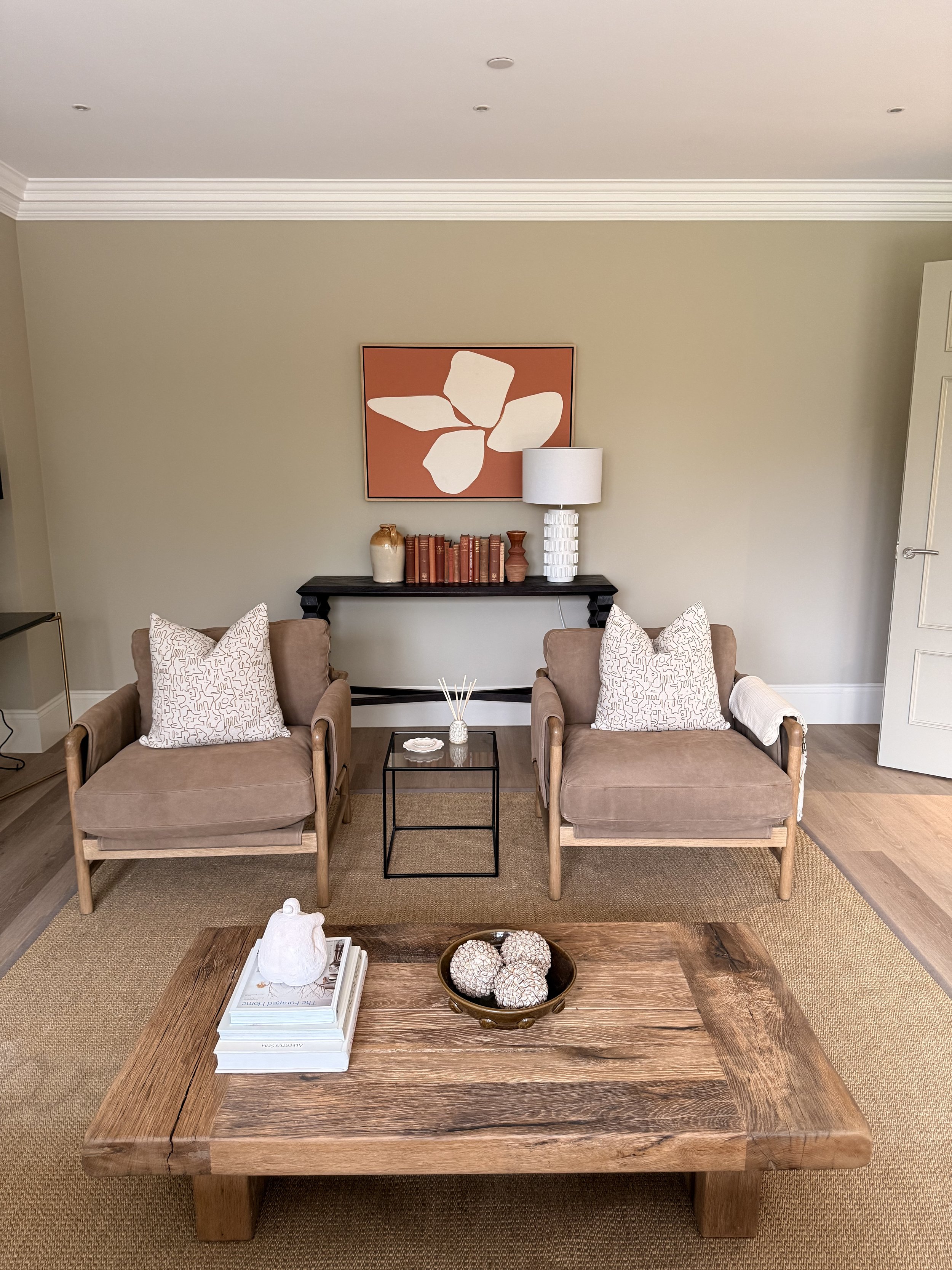 Living room with two beige armchairs with patterned pillows, a large wooden coffee table with decorative objects and books, a black console table with lamps and books against the beige wall, and a framed abstract artwork above the console, with a carpeted floor.