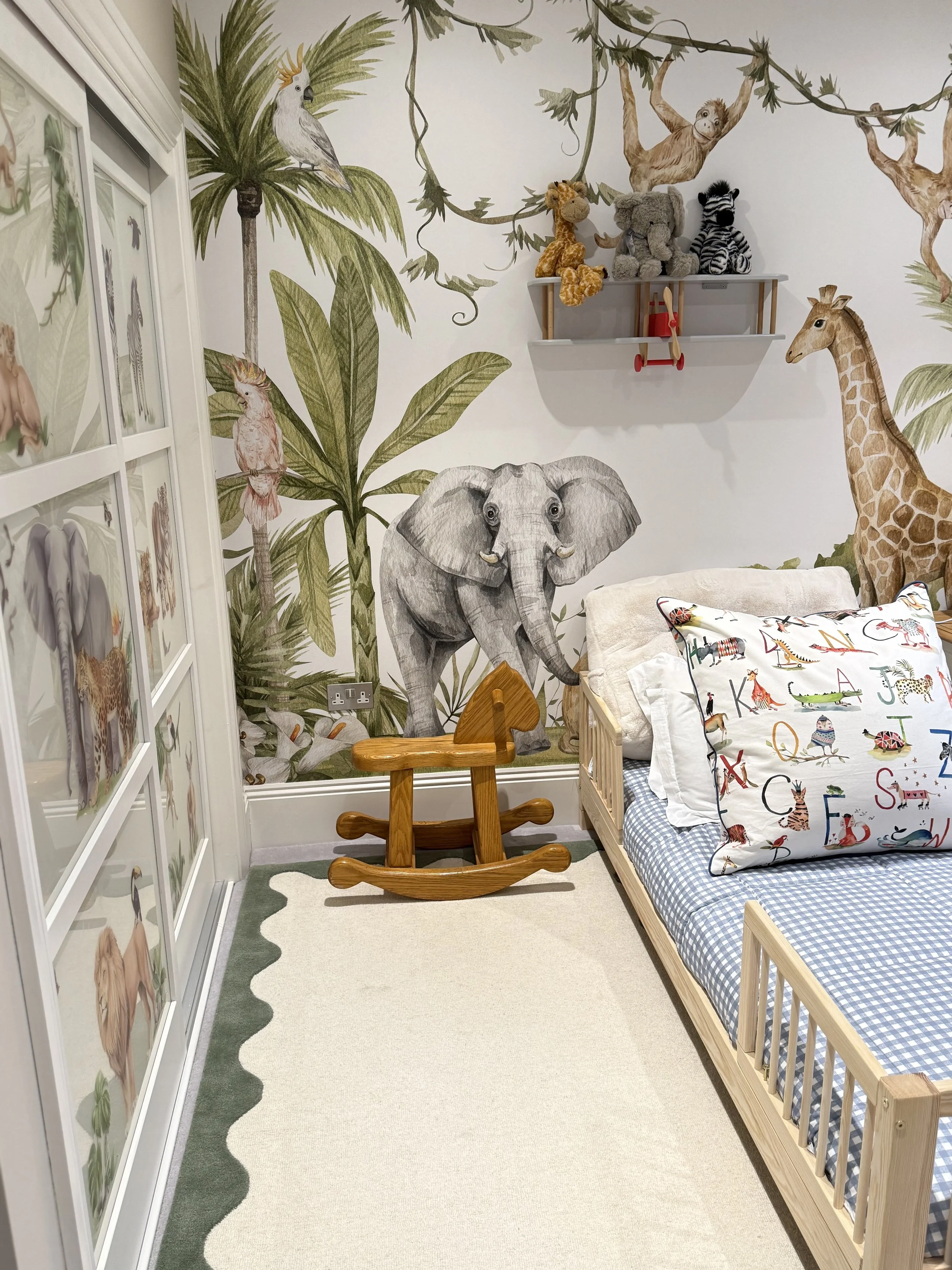 Children's bedroom with jungle animal theme wallpaper including illustrations of a giraffe, elephant, zebra, monkey, cockatoo, and bird on leafy trees. Contains a wooden bed with a blue and white checkered mattress, a pillow with colorful animal alphabet design, a small wooden rocking horse, and a wall-mounted shelf with teddy bears and a toy airplane.